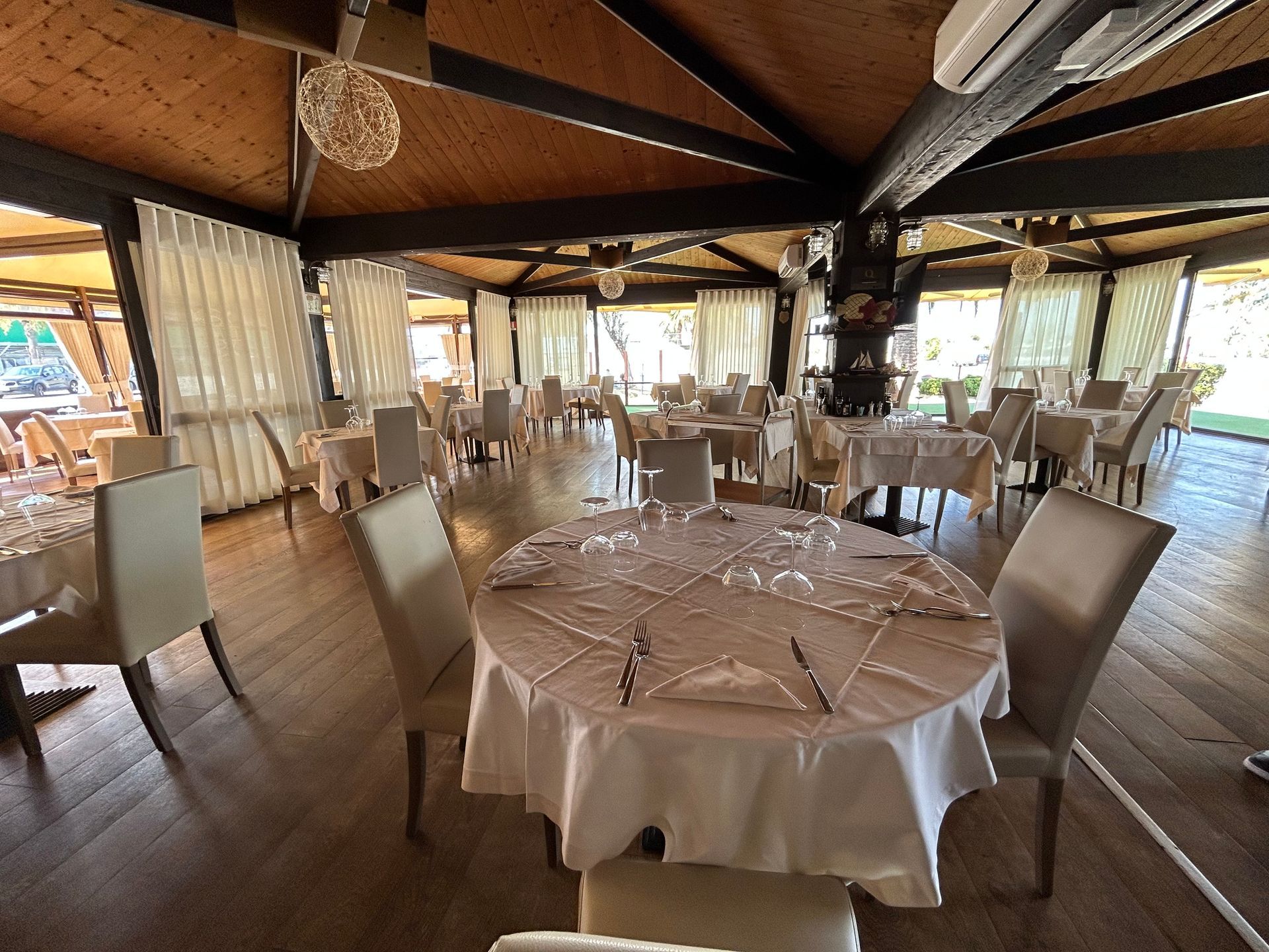 Vista interna di un ristorante. Tavoli rotondi con tovaglie bianche, sedie color crema e ampie finestre.