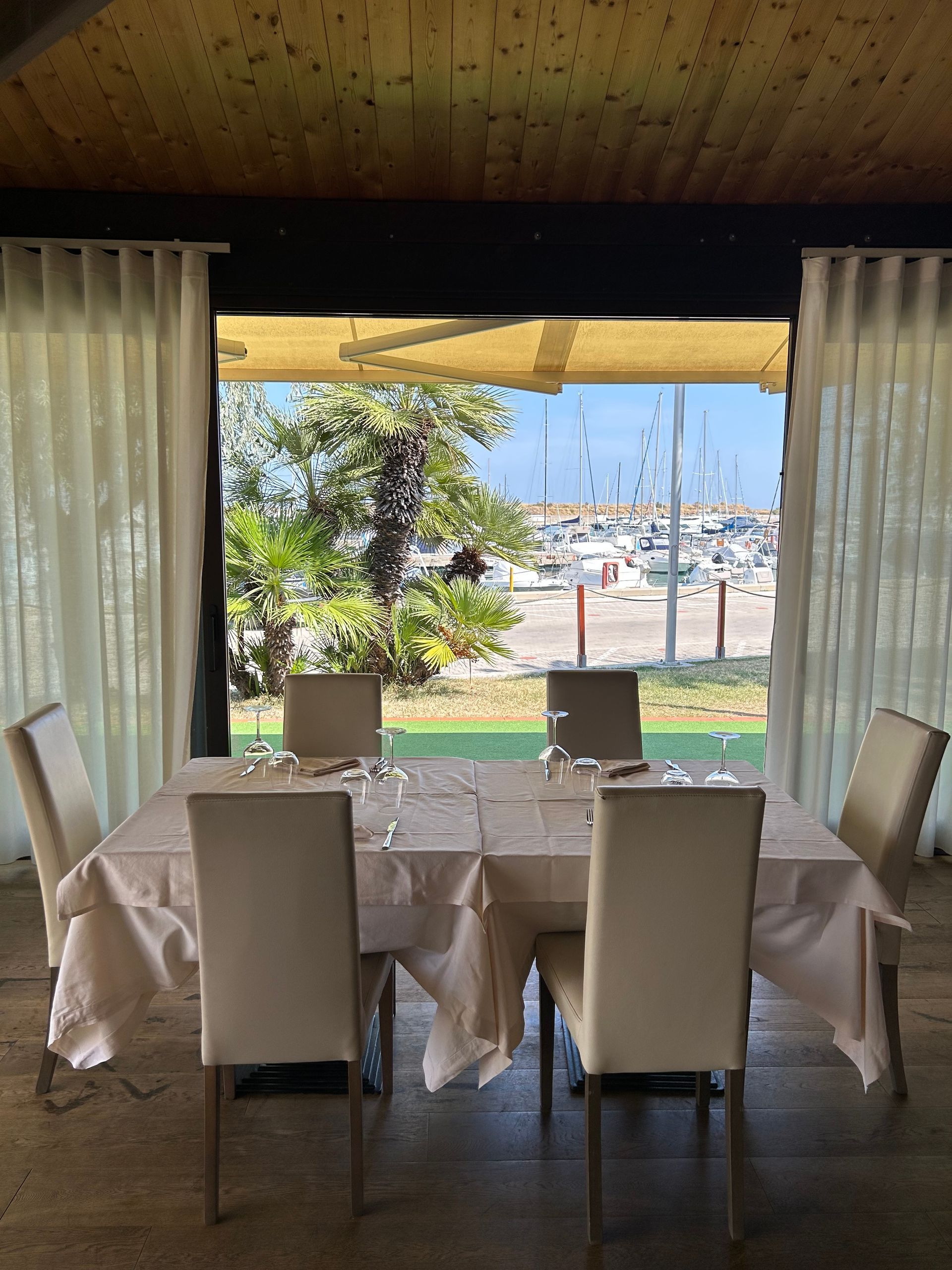 Tavolo da pranzo apparecchiato per sei persone con vista sul porto, sedie color crema e tende bianche.
