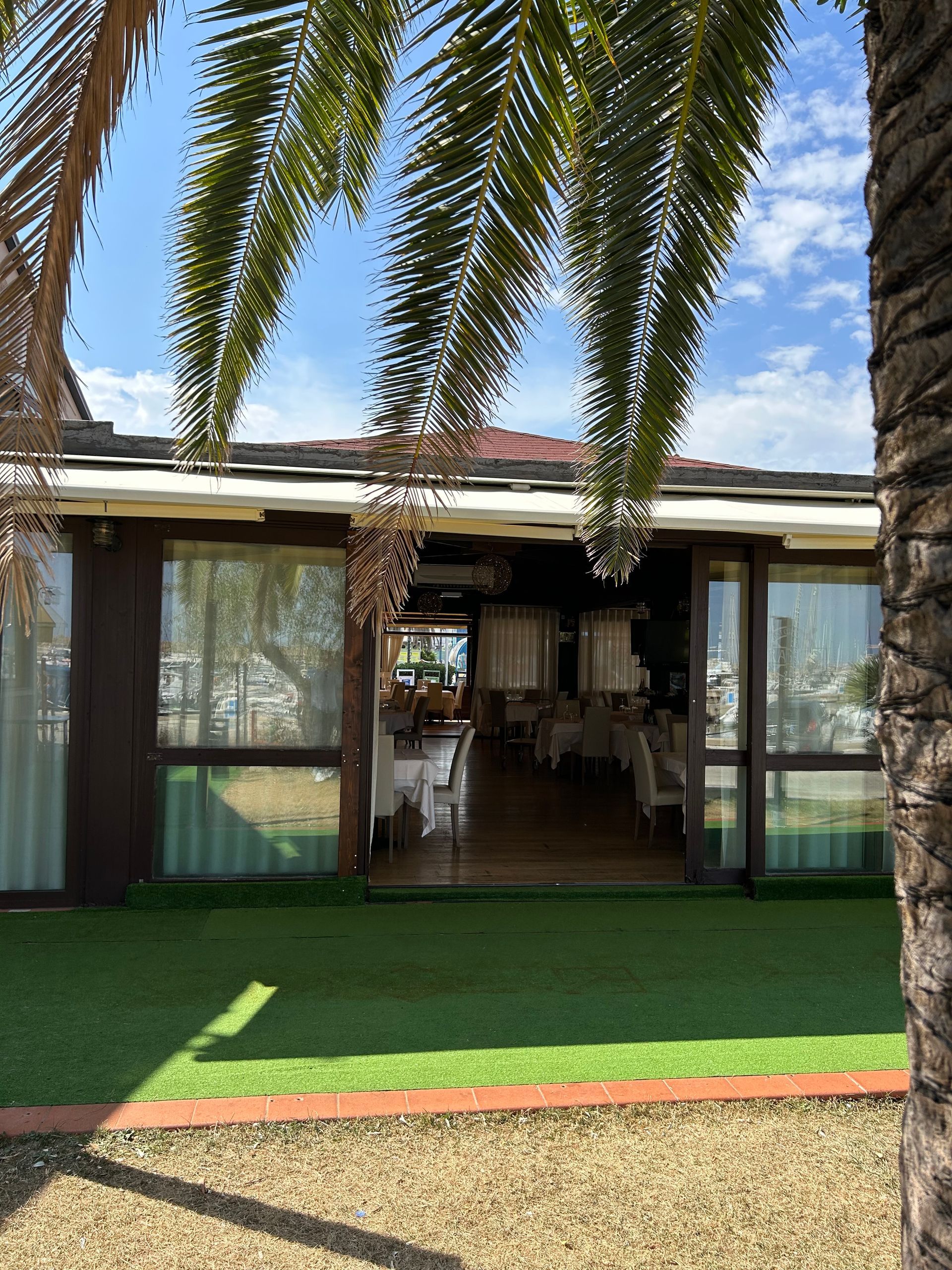 Ristorante con porte a vetri aperte, tavoli apparecchiati all'interno, foglie di palma e cielo azzurro sopra la testa.