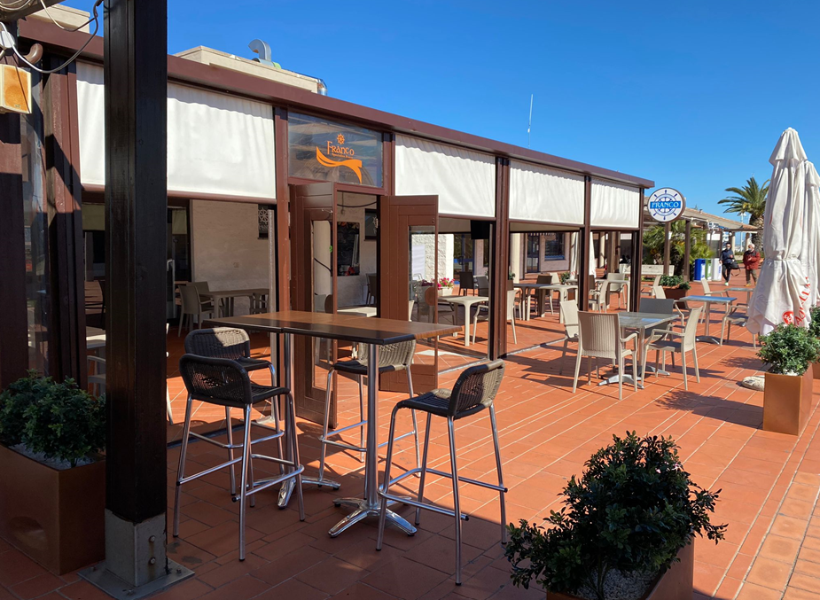 Patio esterno del ristorante con tavoli, sedie e tende da sole. Edificio marrone e bianco. Giornata di sole.