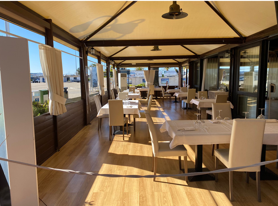 Patio del ristorante con tavoli apparecchiati per cenare, pavimenti in legno, tende da sole color cuoio e ampie finestre.