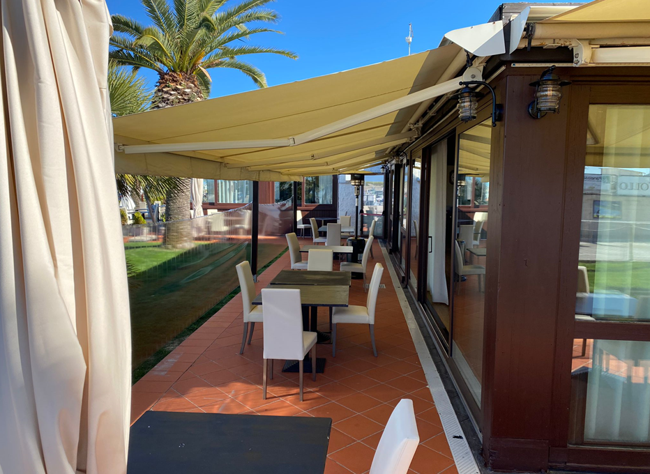 Posti a sedere all'aperto del ristorante con tenda da sole, tavoli e sedie, accanto a un edificio e al prato.