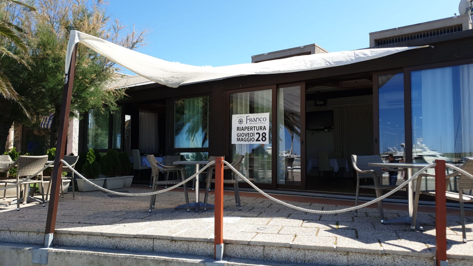 Ristorante sulla spiaggia con tenda da sole, porte a vetri e tavoli. Un cartello appeso alla finestra. Barriera di corda.