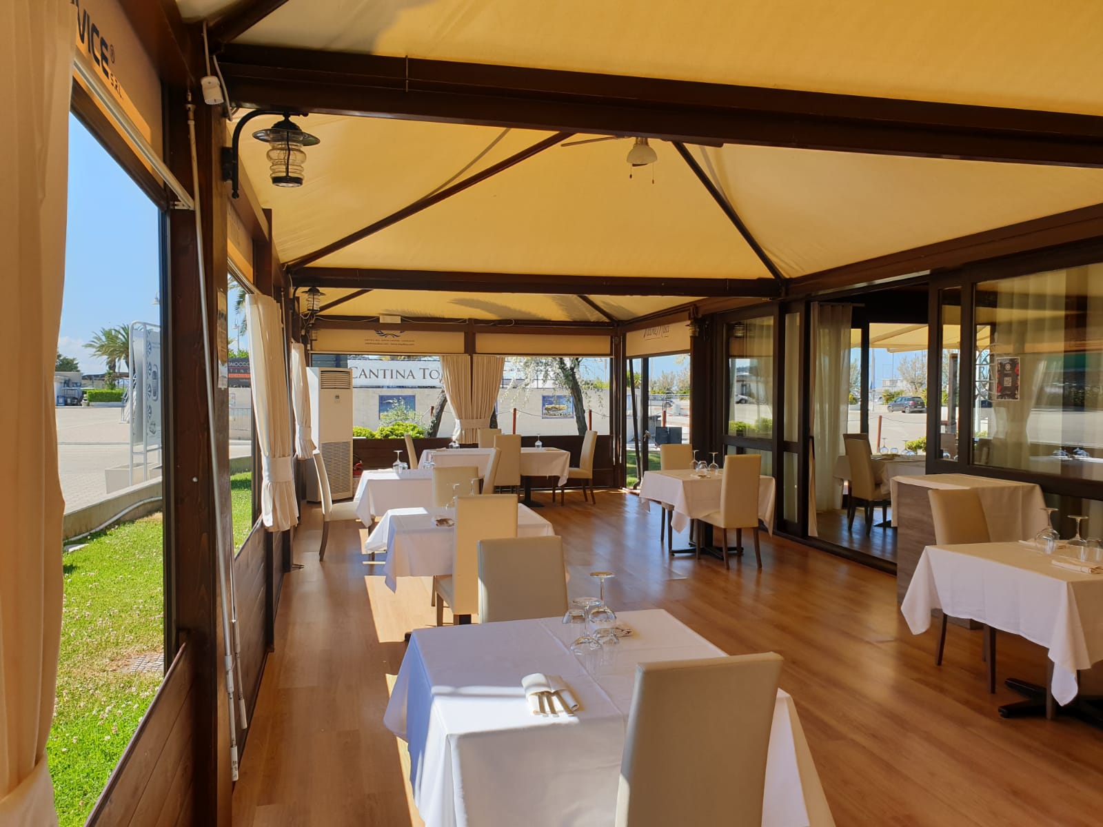 Ristorante con tovaglie bianche, tende color cuoio, travi in ​​legno, vetrate e vista soleggiata.