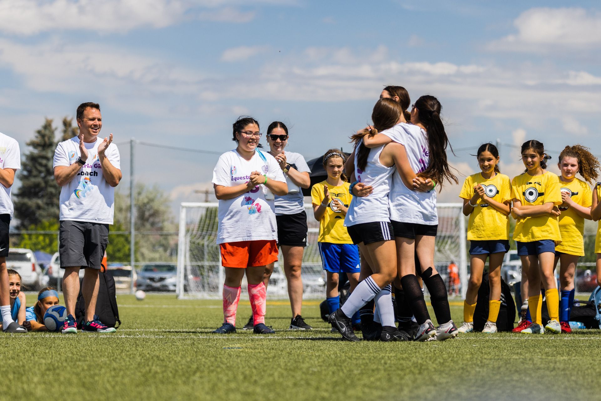 Soccer players hugging on a field, others cheering. Sun, blue sky, green grass.