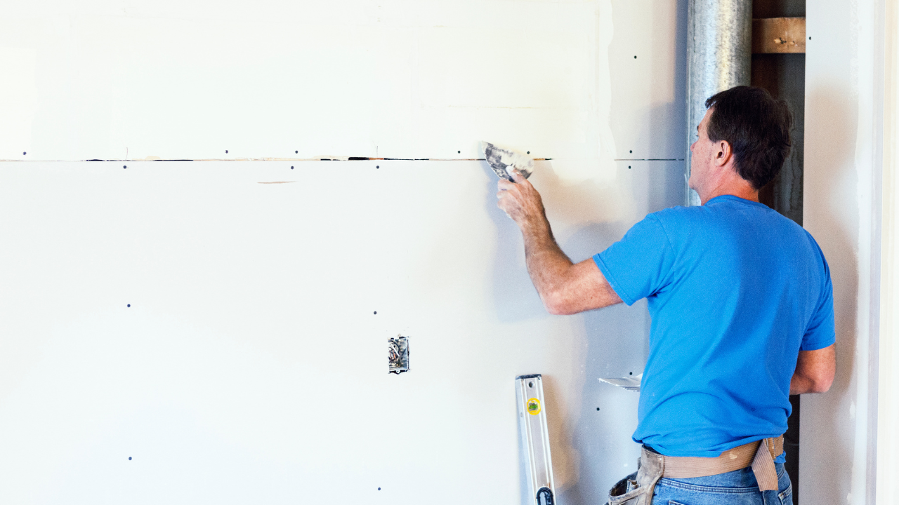 Man in blue shirt patching drywall with a trowel in a white room near a ladder