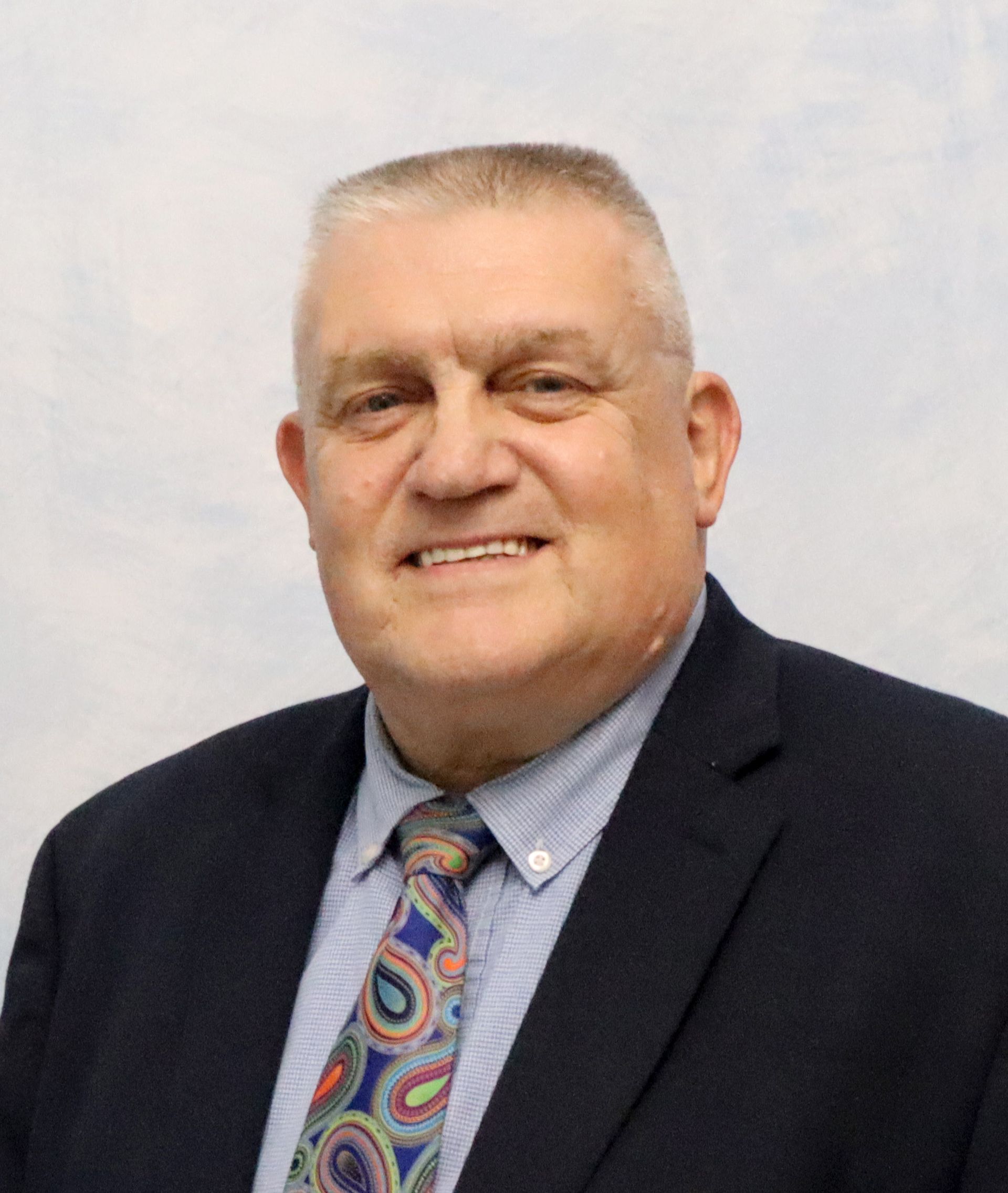 Man in glasses and suit smiles at camera. Gray background.