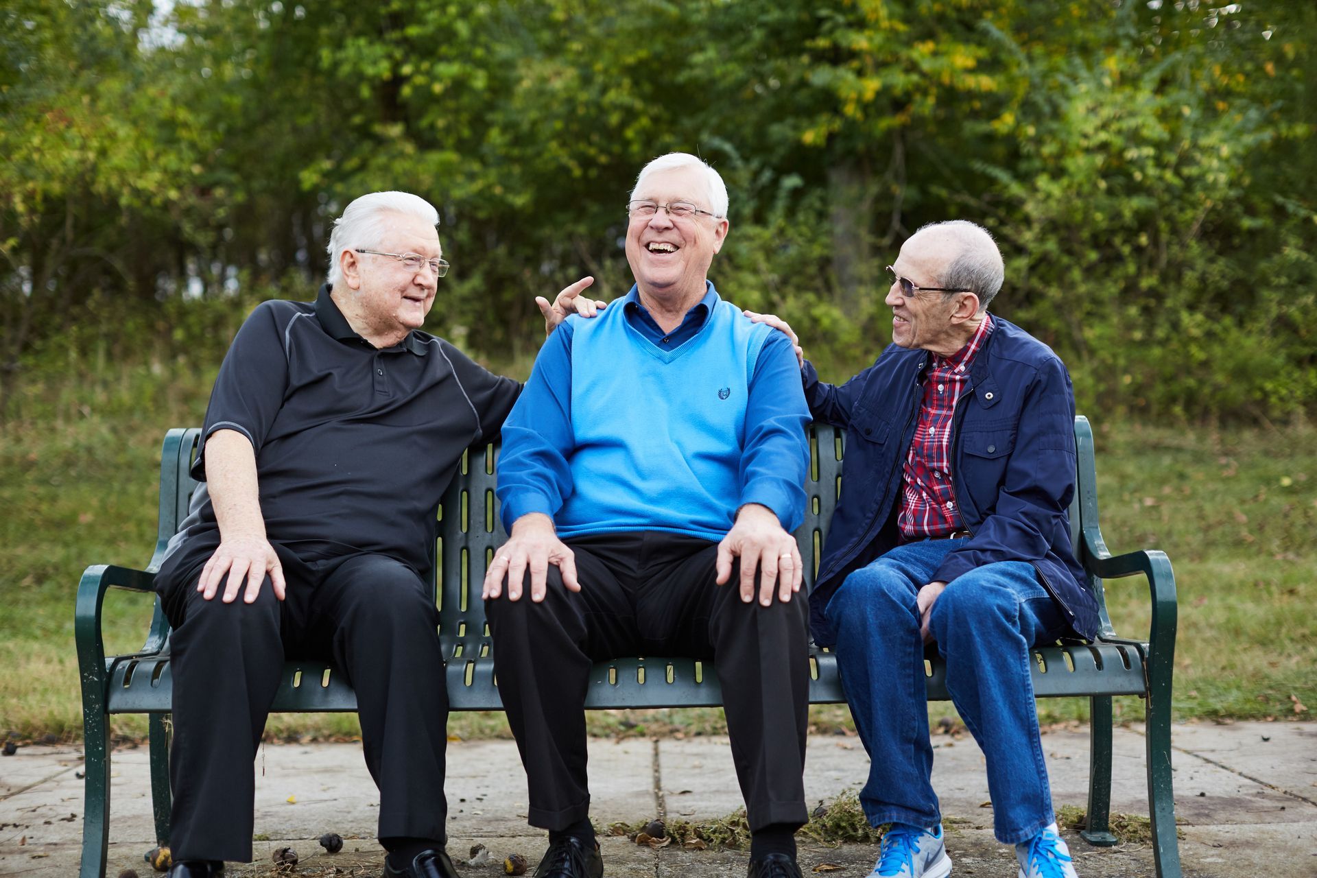 Three older men laughing on a park bench outdoors. One has his arm around another's shoulder.