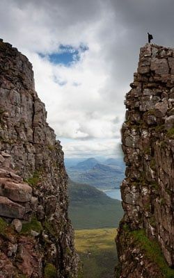 A person stands on a rocky mountain peak, overlooking a valley and lake under a cloudy sky.