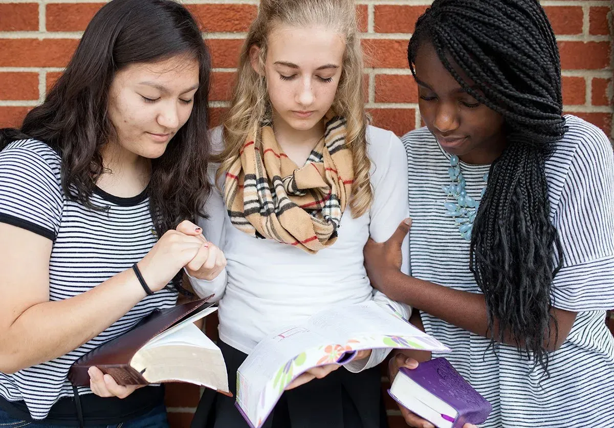 Three diverse young women reading and smiling together on outdoor steps.