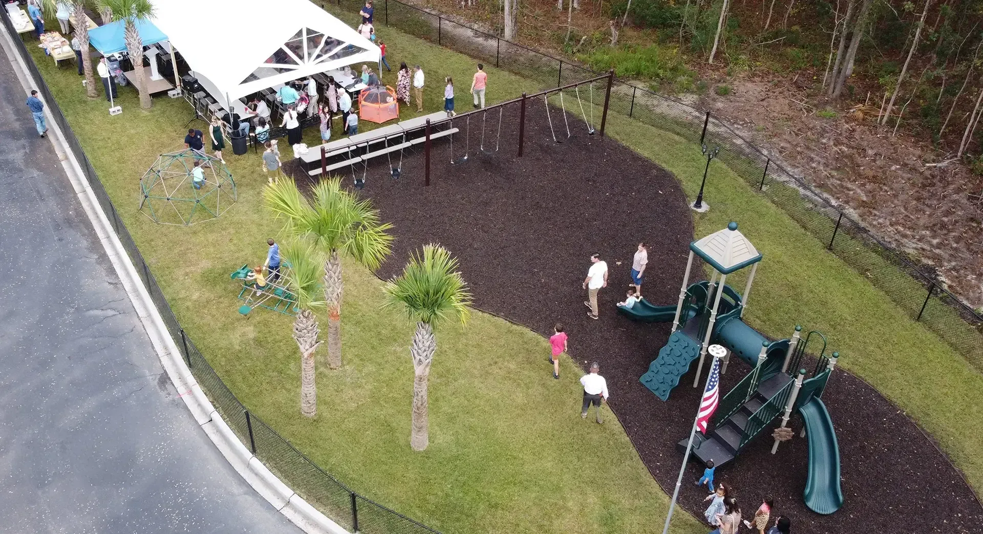 Overhead view of playground, party tent, and people. Palm trees dot the grassy area.