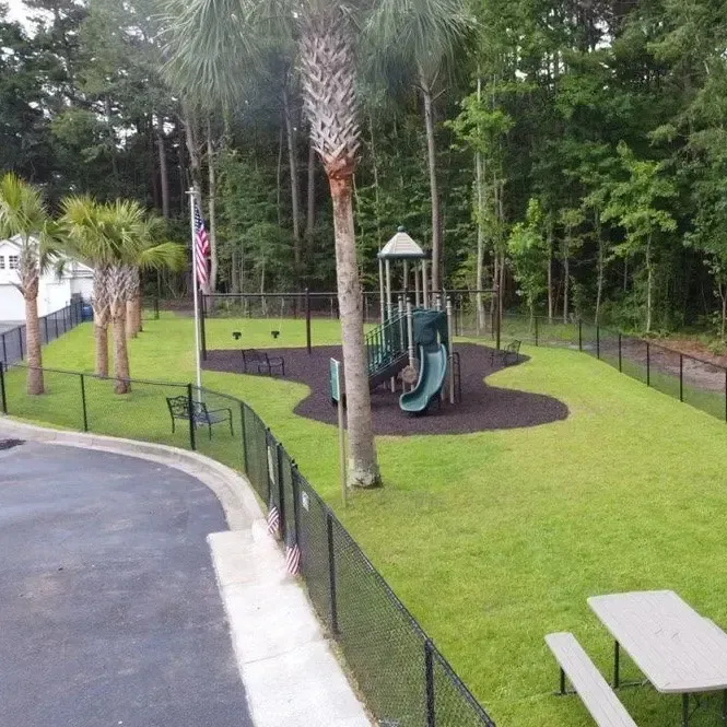 A fenced-in playground with a slide, swings, and picnic table, surrounded by green grass and trees.