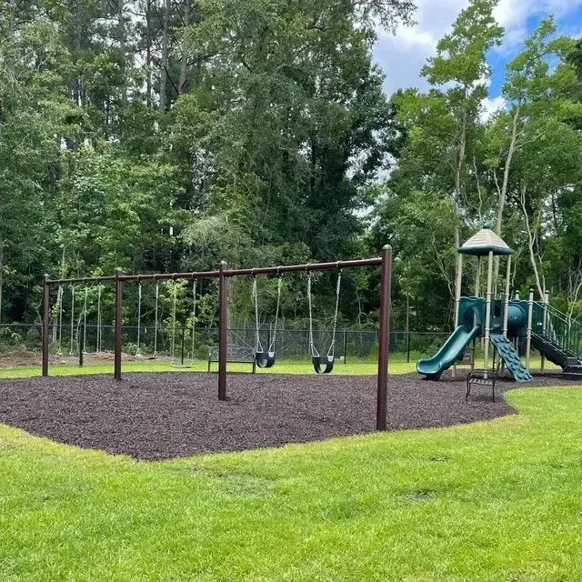 Playground with swings and slide on a bed of mulch surrounded by green grass, trees, and a blue sky.