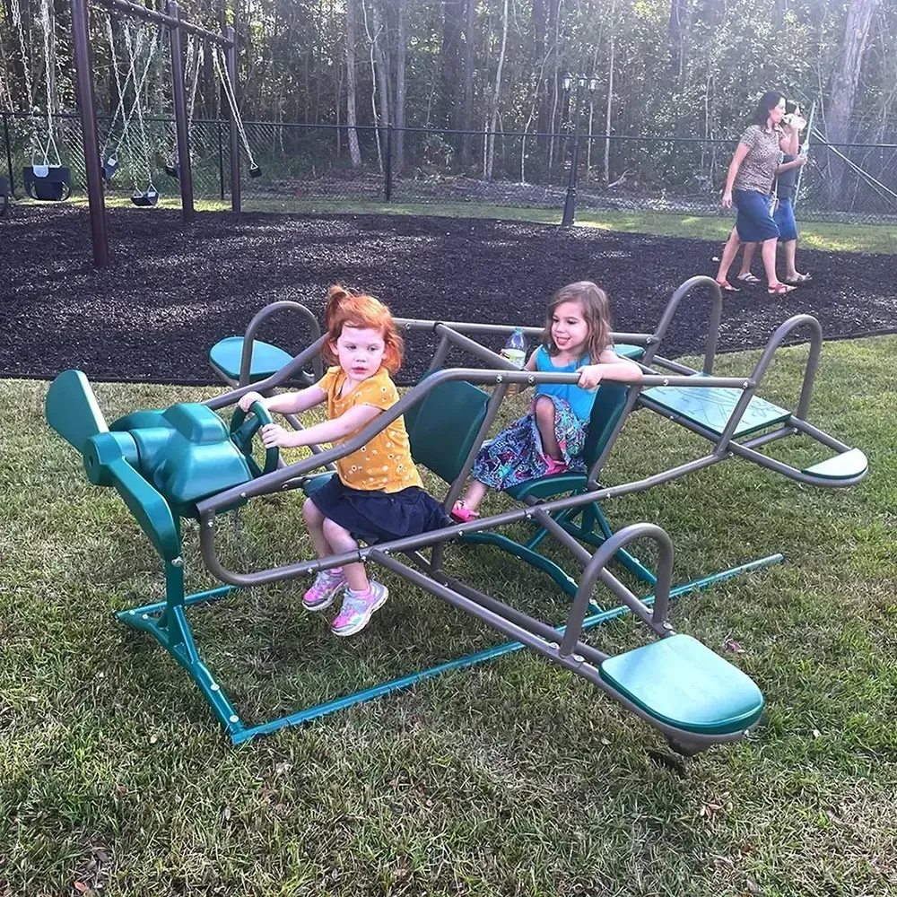 Two girls on a green airplane seesaw at a playground; trees and adults in the background.