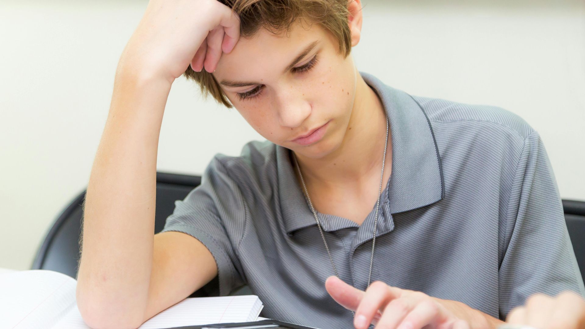 Young man in a gray shirt studying at a desk, resting his head on his hand with a focused expression
