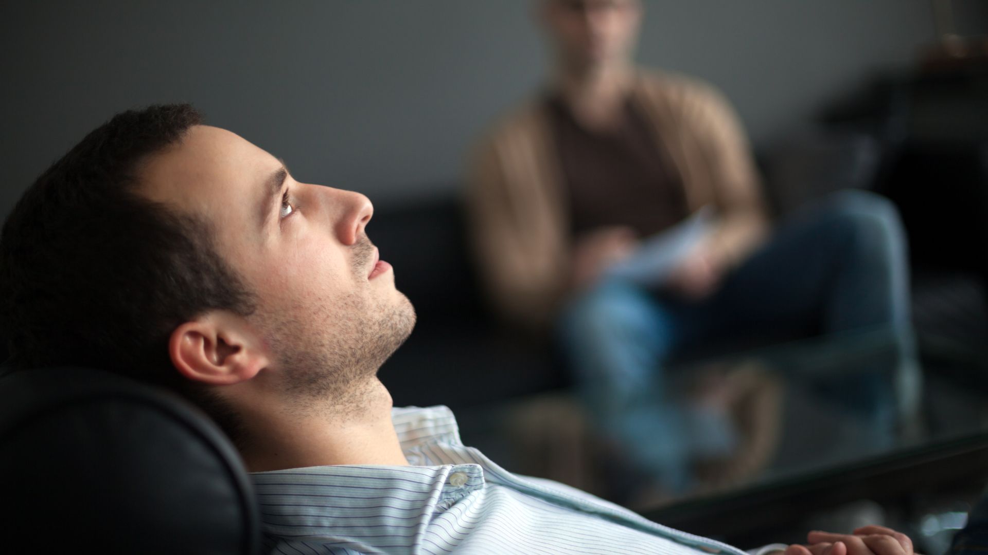 Man reclining on a couch while a blurred person sits in the background, suggesting a counseling session