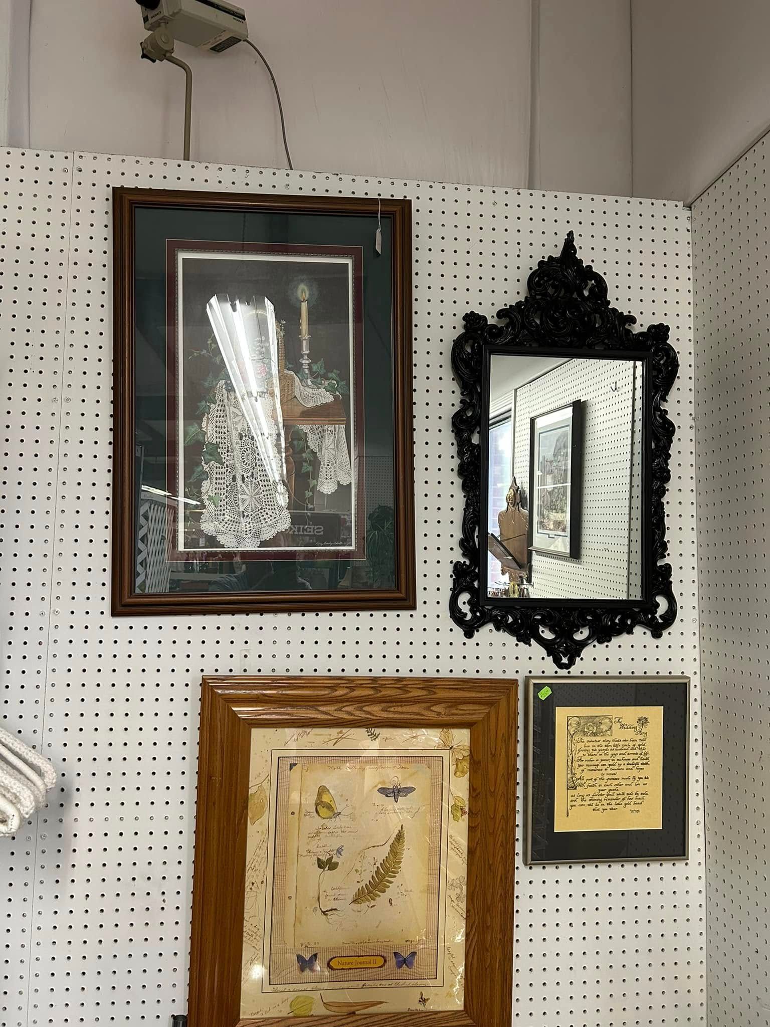 Three framed artworks and a black ornate mirror on a pegboard wall.