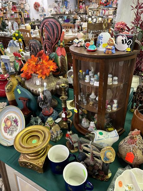 Cluttered antique shop display with ceramics, glassware, flowers, and figurines on shelves and tables