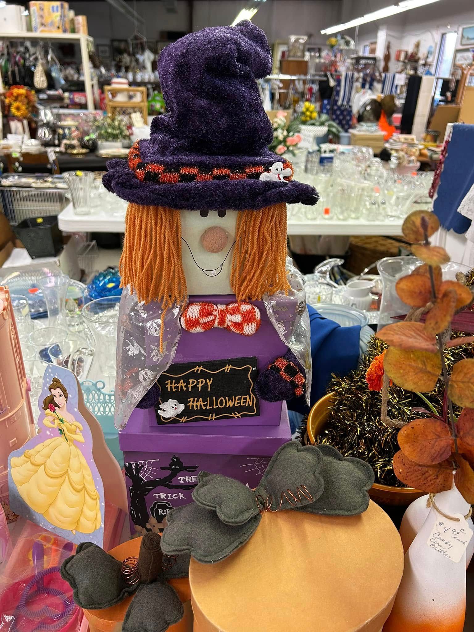 Handmade witch scarecrow decoration with purple hat, orange hair, and pumpkins at a craft shop display