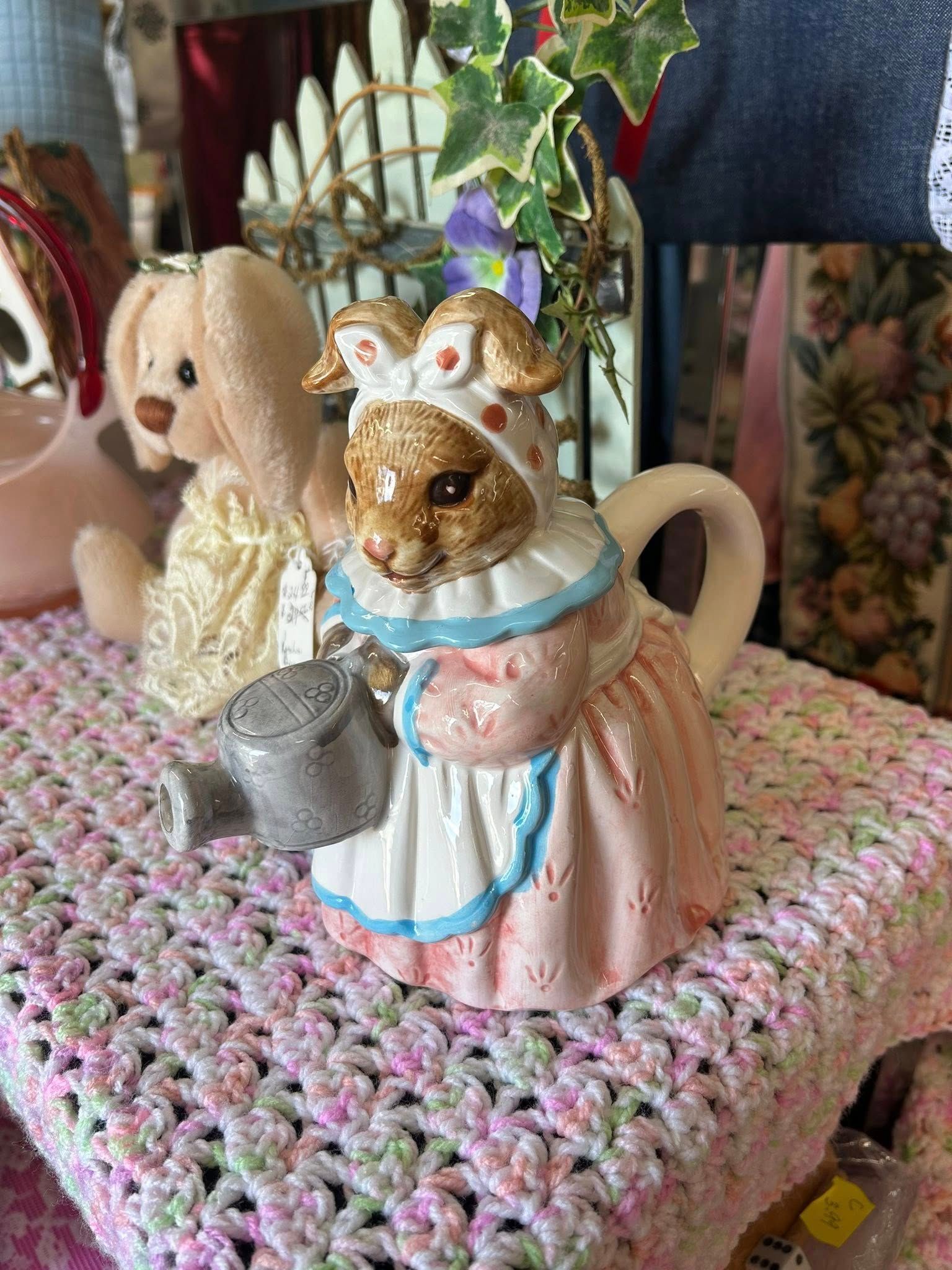Two small dog figurines in pastel dresses on a pink crocheted mat with a teapot and flowers behind them