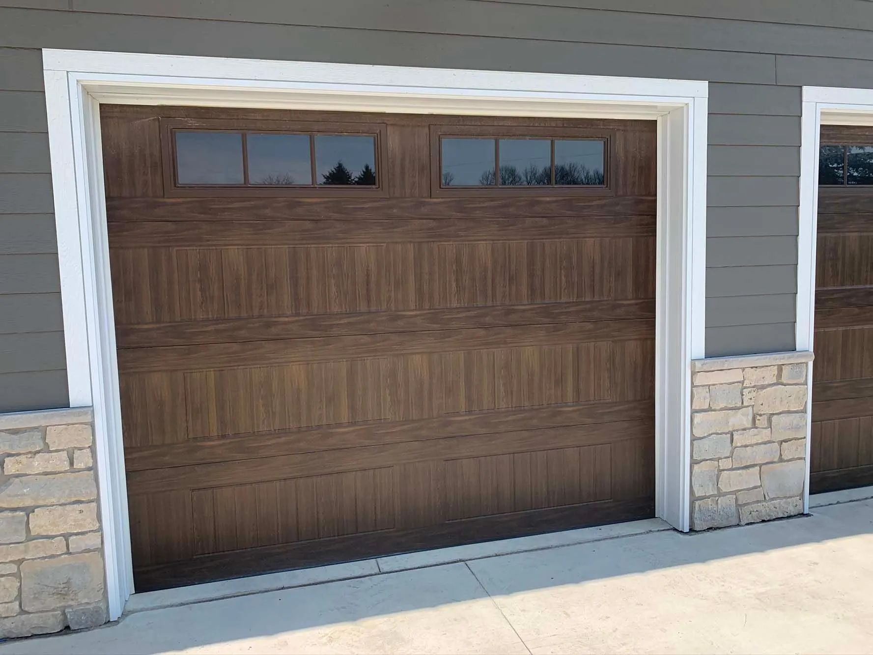 Wood-paneled garage door with small windows, framed by white trim and stone siding on a driveway