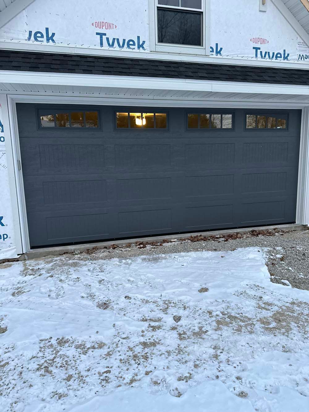 Gray garage door with four small window panels at the top, covered in Tyvek, snow on the ground.