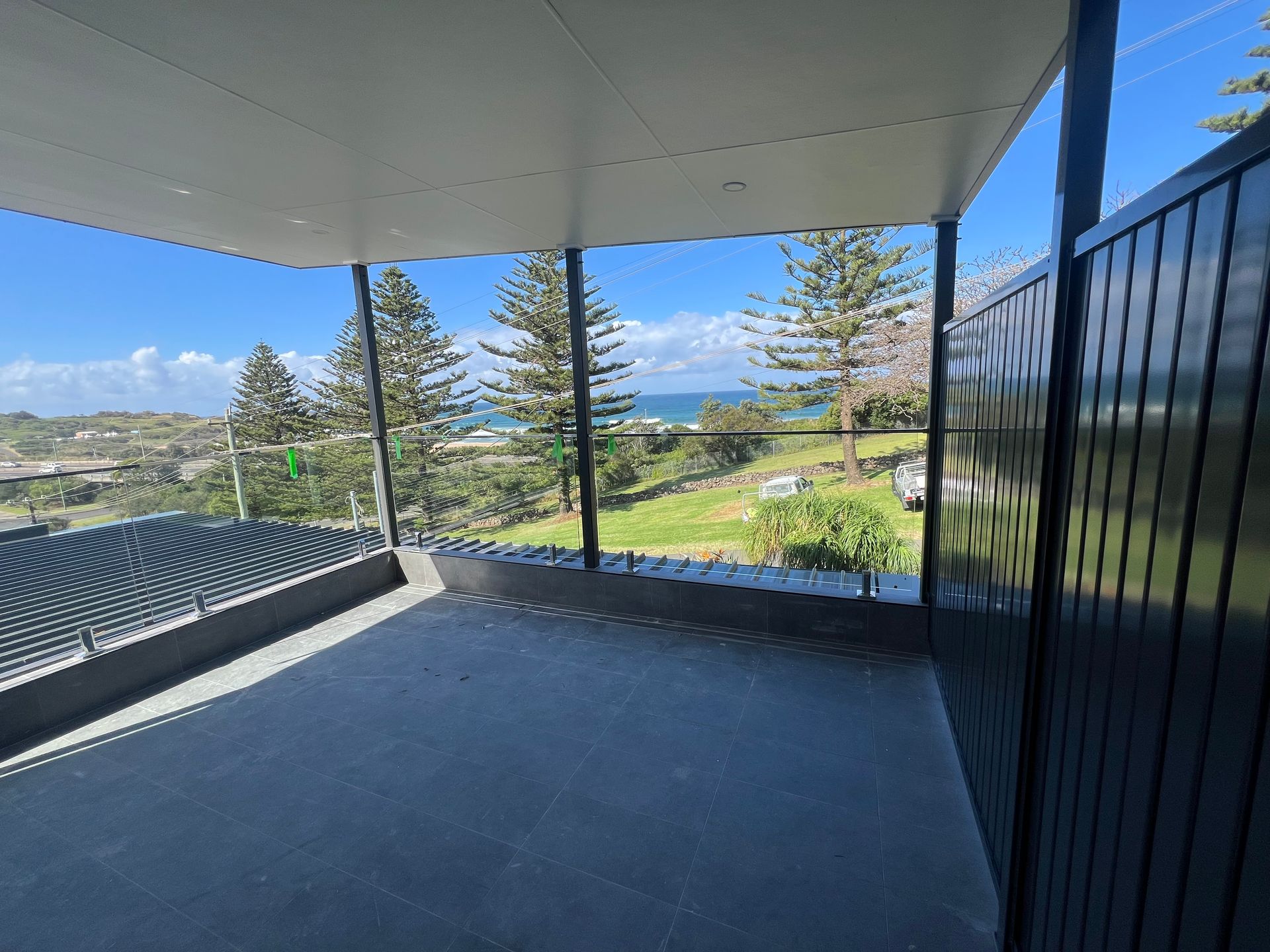 Covered patio with blue floor, black railing, and ocean view beyond the yard.  — Taddeo Constructions in Kiama Downs, NSW