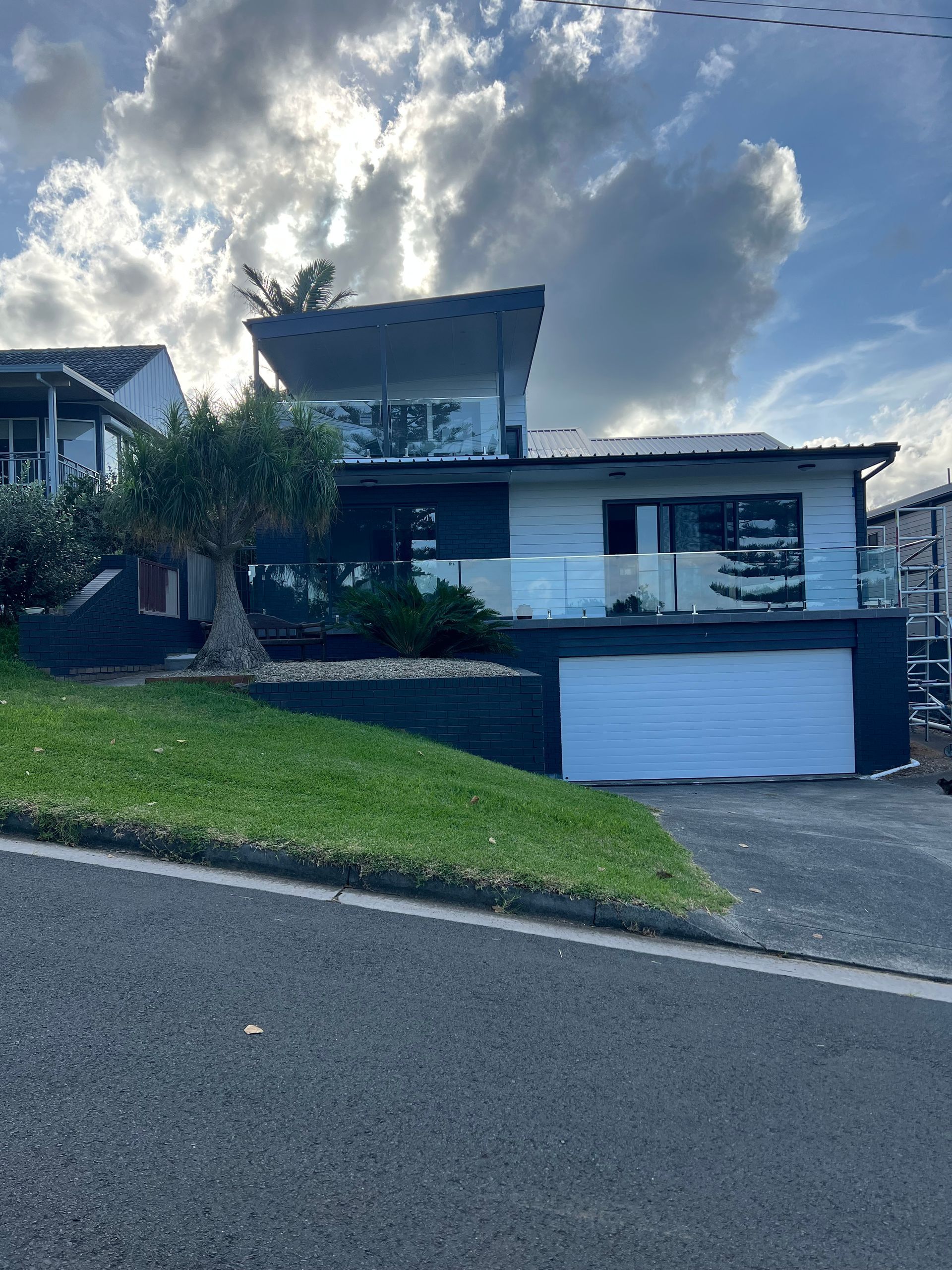 Modern house on a sloped street under a cloudy sky, with a garage and green lawn.  — Taddeo Constructions in Kiama Downs, NSW