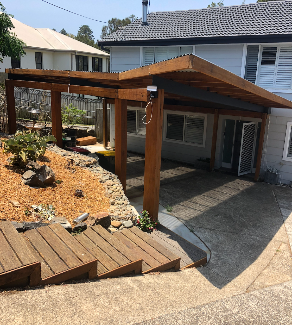 Wooden pergola and covered patio beside a house, with gravel landscaping and steps on a sunny day