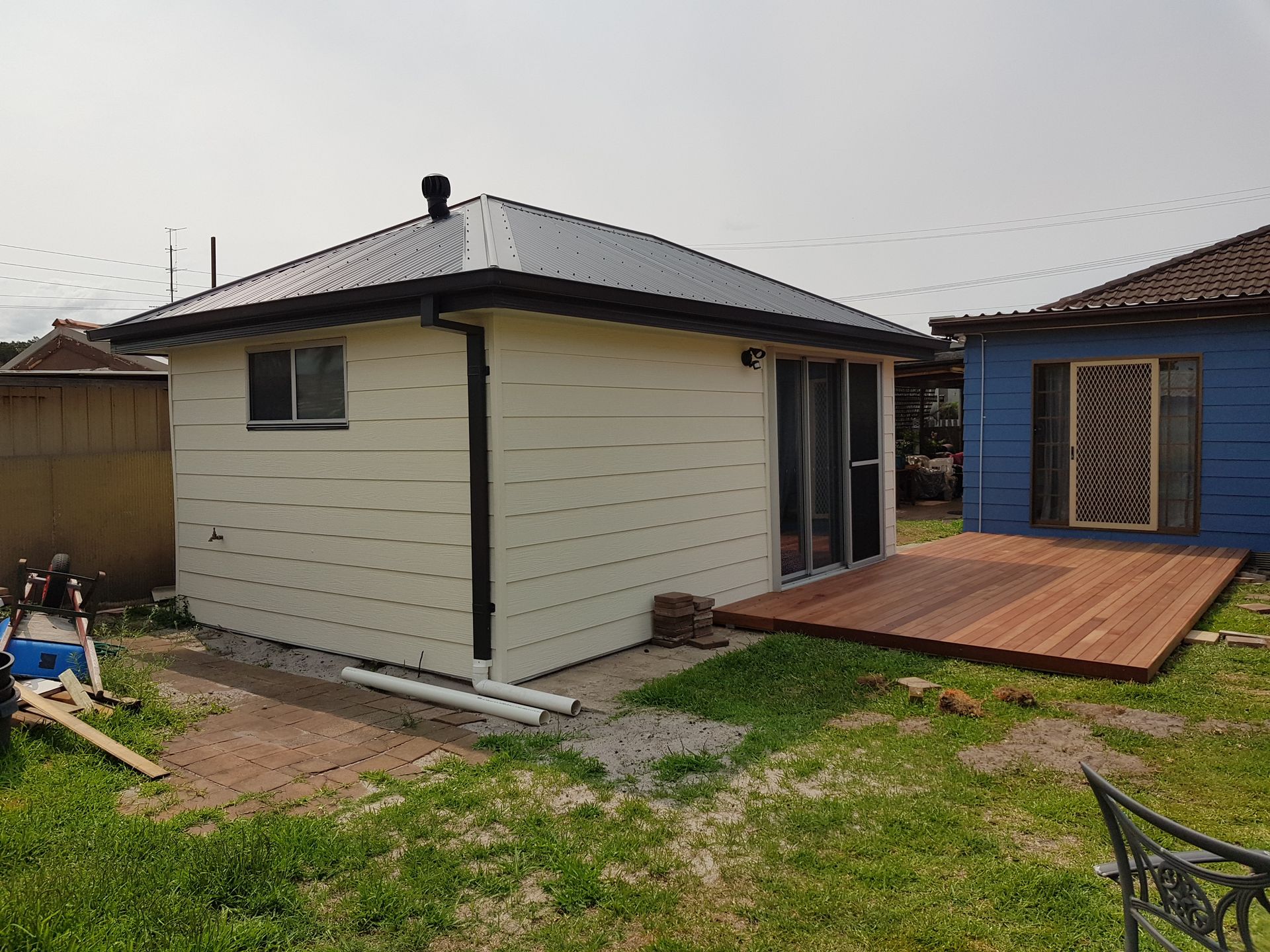 Small beige shed with dark roof and wooden deck in a grassy backyard, beside a blue garage.  — Taddeo Constructions in Kiama Downs, NSW