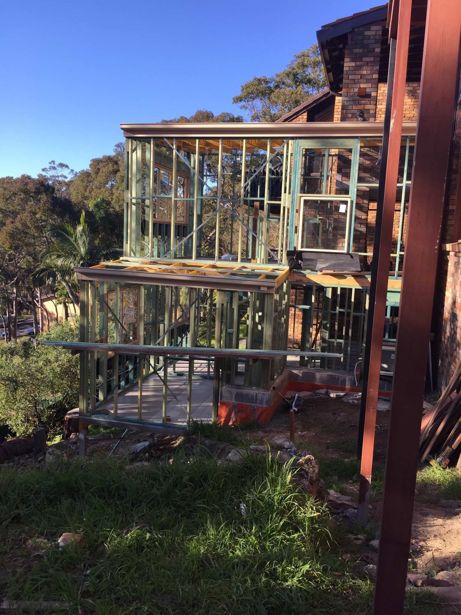 Partially built wooden deck and frame beside a brick house, surrounded by trees and grass.