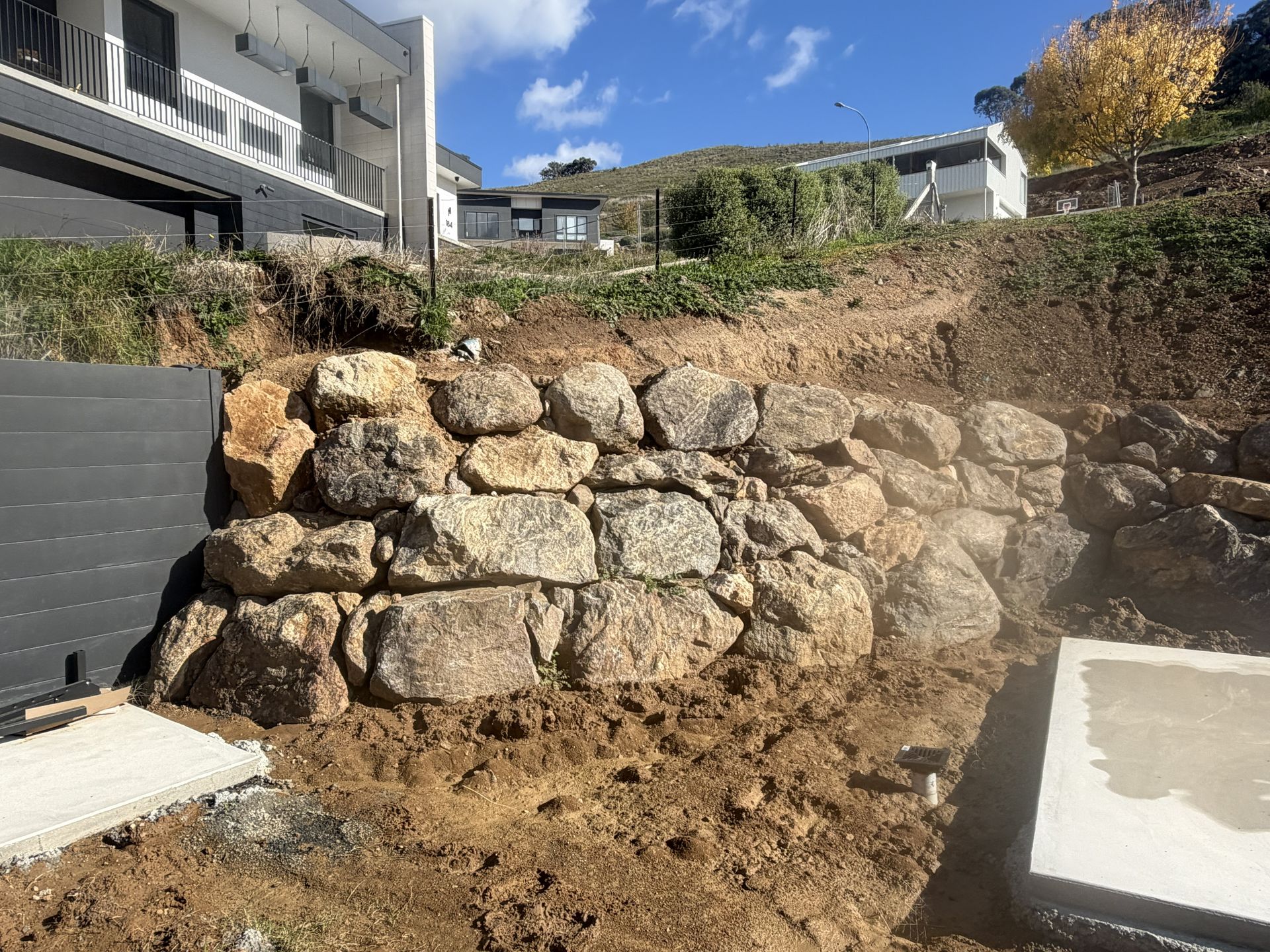 Stone retaining wall under construction in a residential yard with dirt, steps, and houses nearby