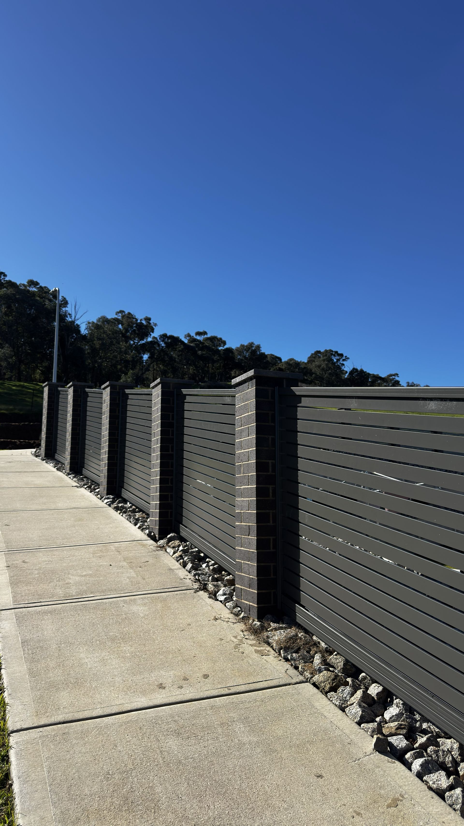 Concrete sidewalk beside dark slatted fence under a clear blue sky