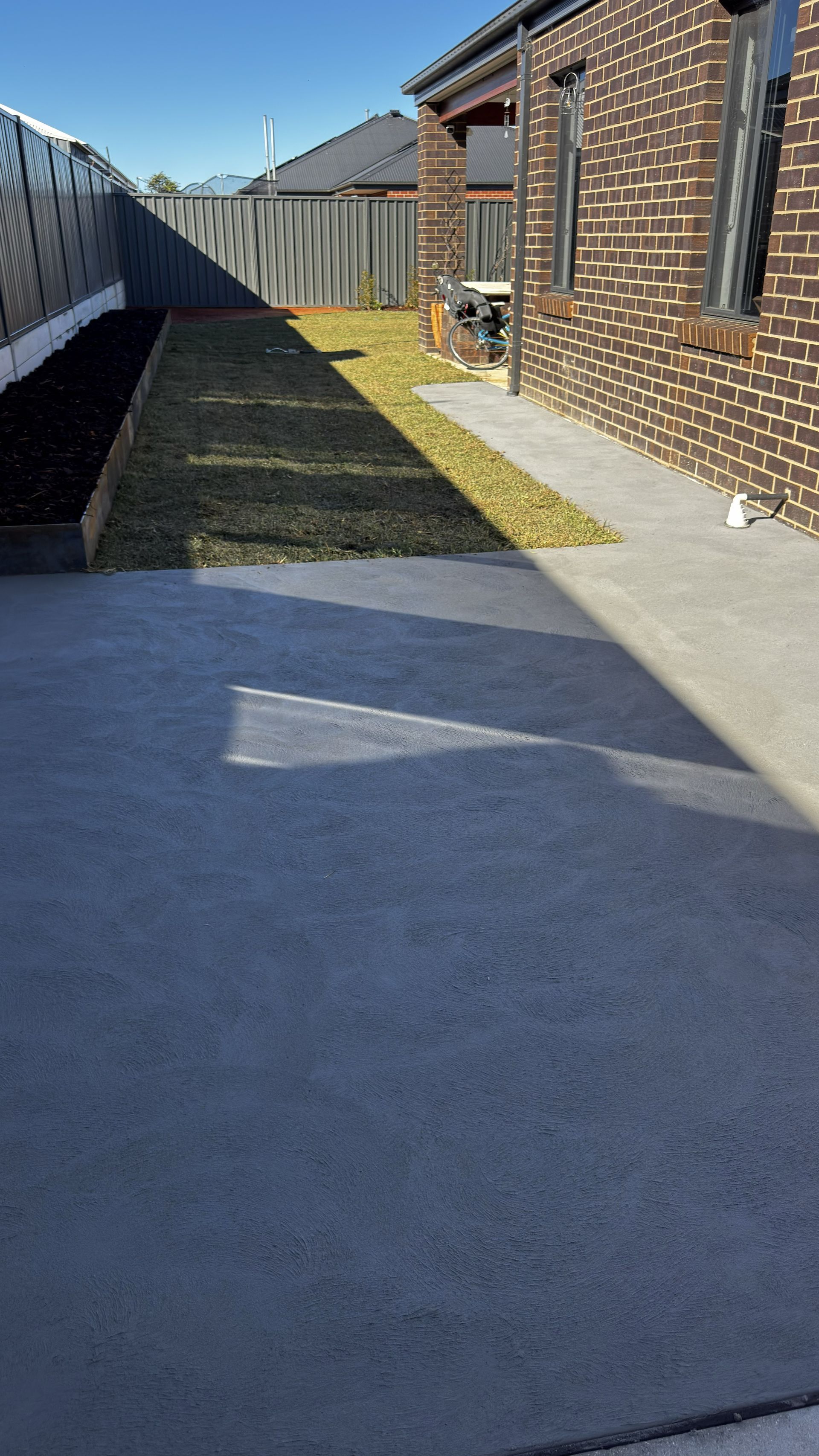 Concrete backyard walkway beside a brick house and fence, with a narrow grassy strip in between.