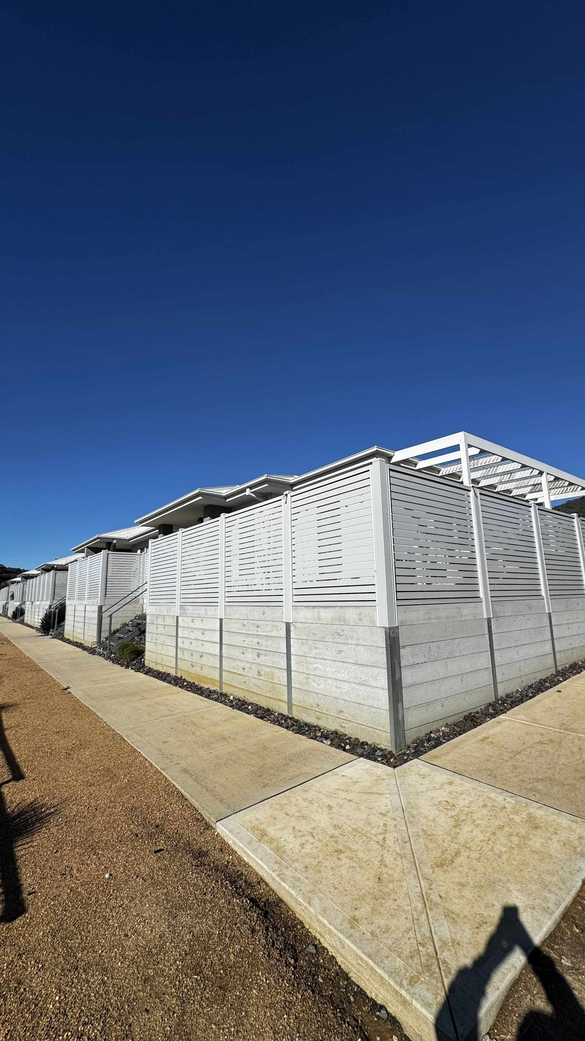 White metal fence around a building corner under a clear blue sky, beside a gravel path.