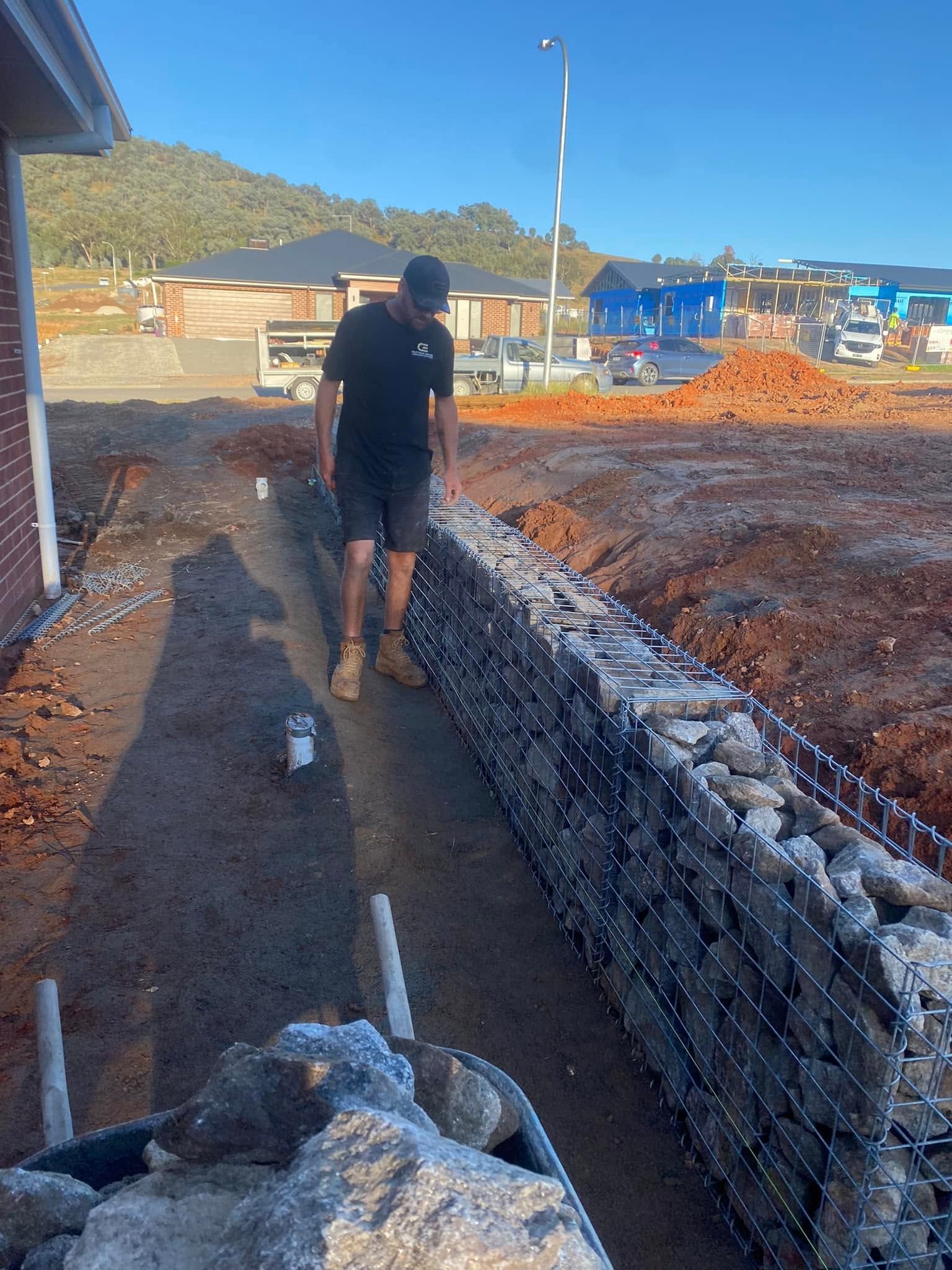 Person walking along a gravel path beside a gabion wall at a construction site under clear sky