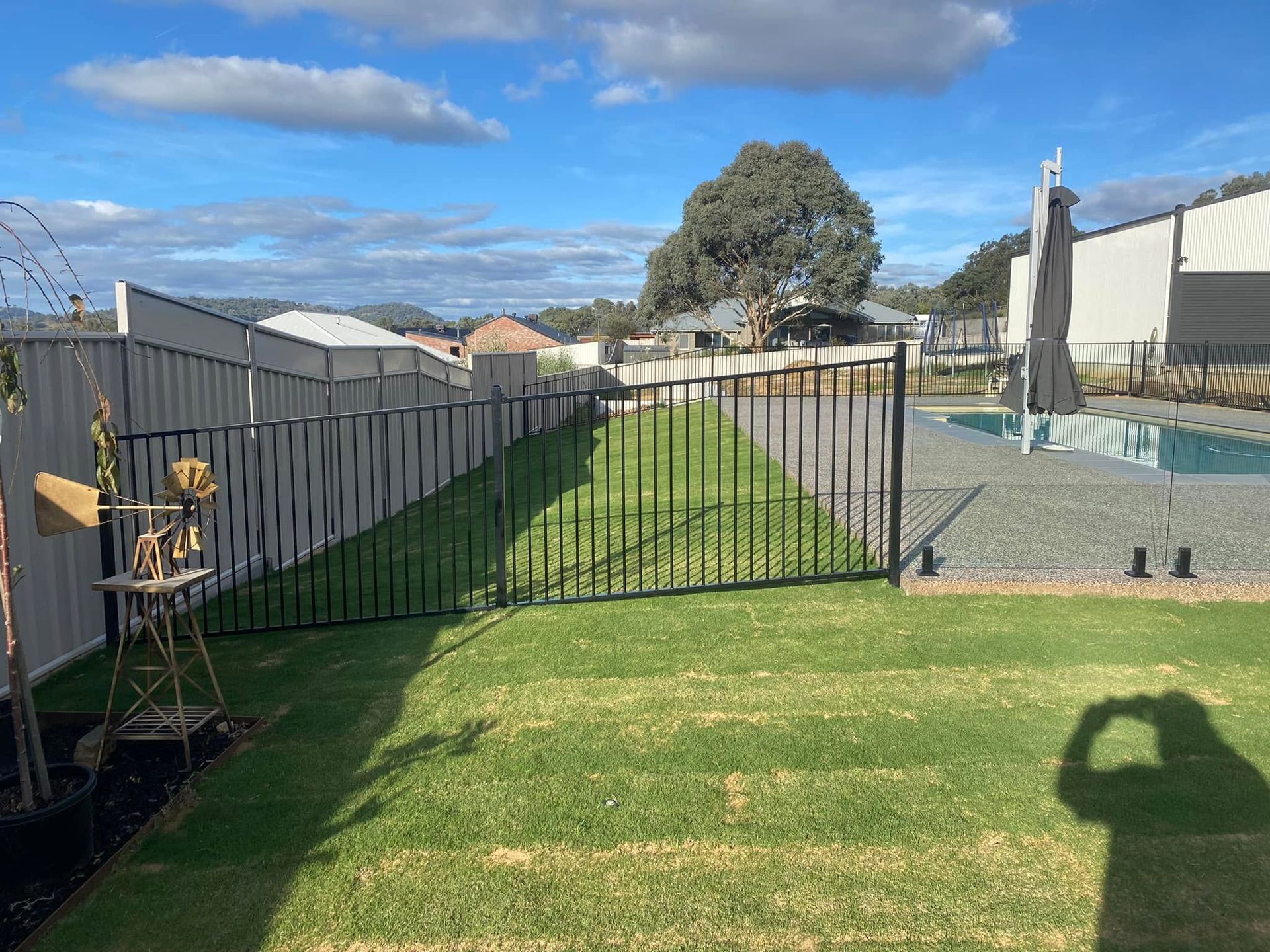 Backyard with green lawn, black fence, tree, and gravel area under a blue sky