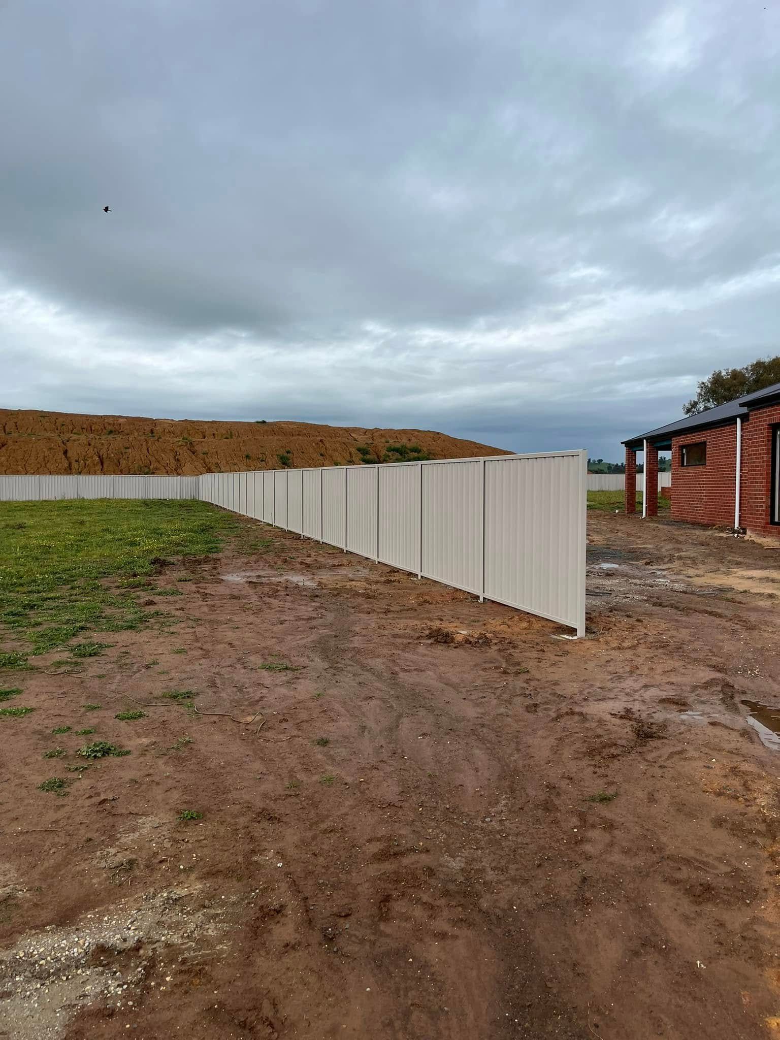 A white corrugated fence lines a muddy dirt lot under a cloudy sky beside a brick building.