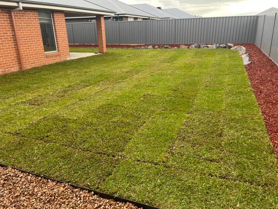 Backyard lawn bordered by brick house, gray fence, and red mulch beds.
