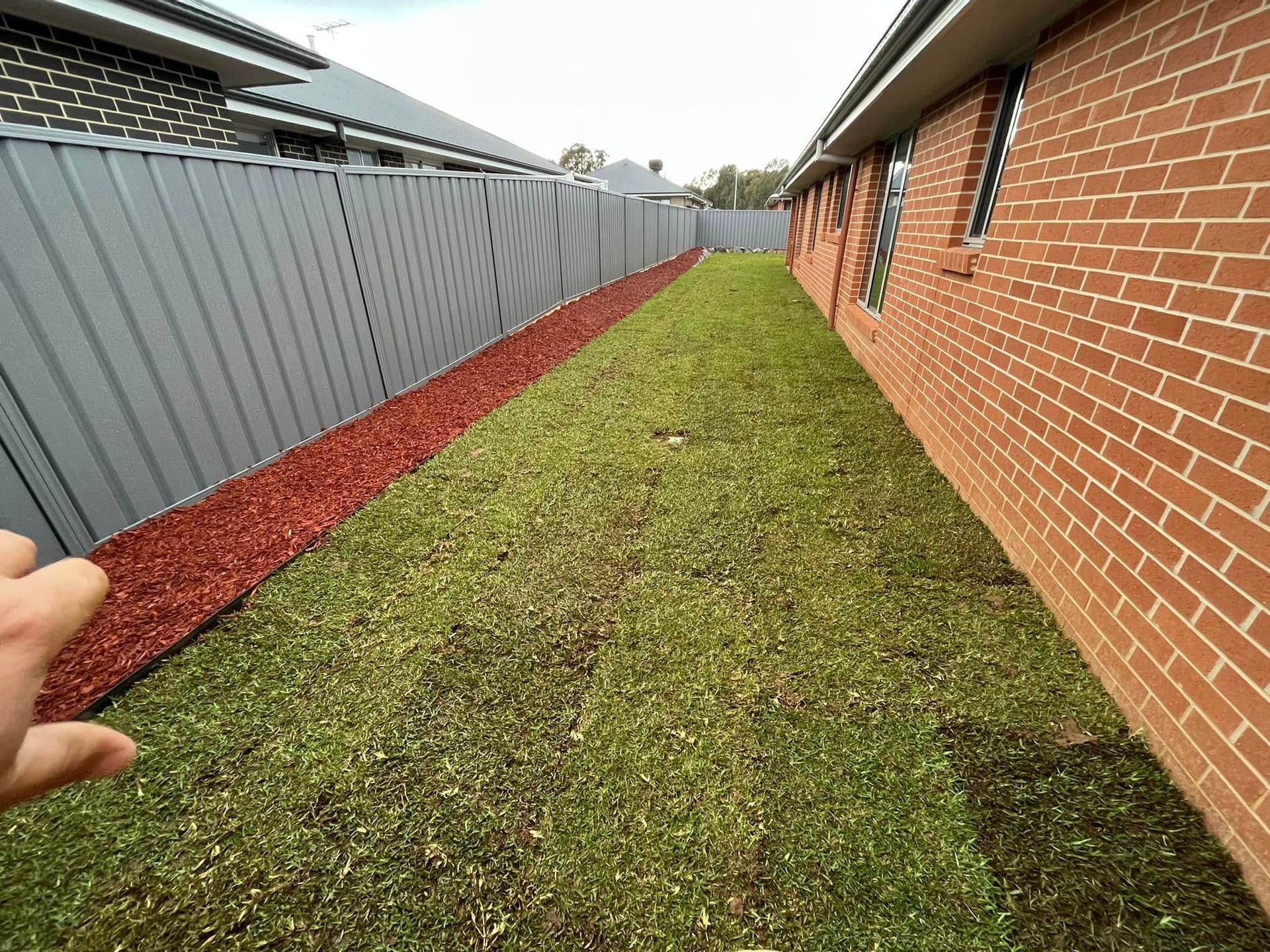Narrow grassy side yard between a brick house and a gray fence with red mulch border