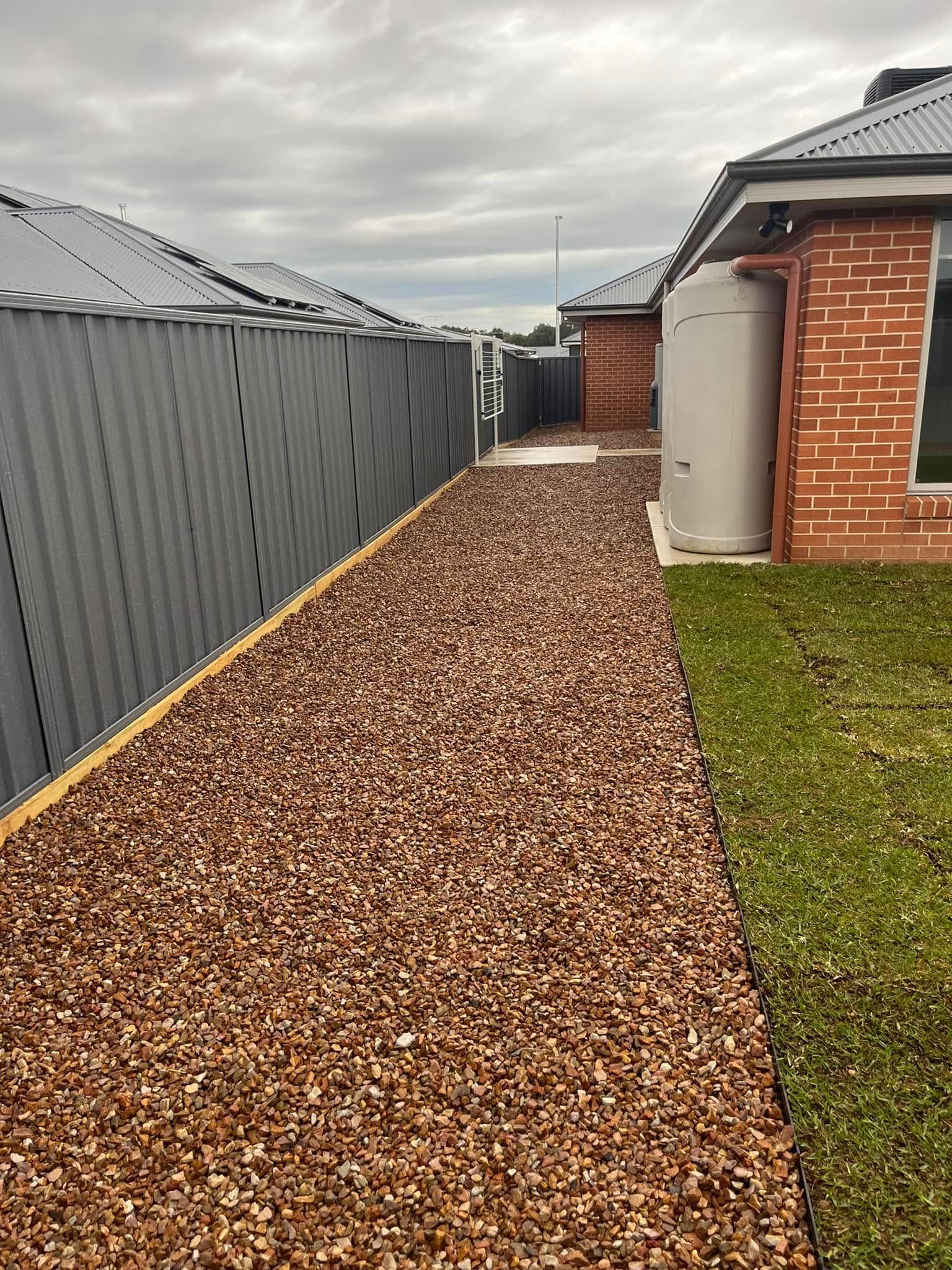 Gravel path beside a gray fence and brick house, leading to a shed in a suburban yard