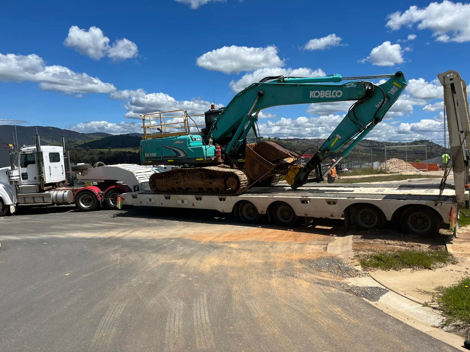 Excavator loaded on a semi-trailer beside a road under a blue sky with mountains in the background