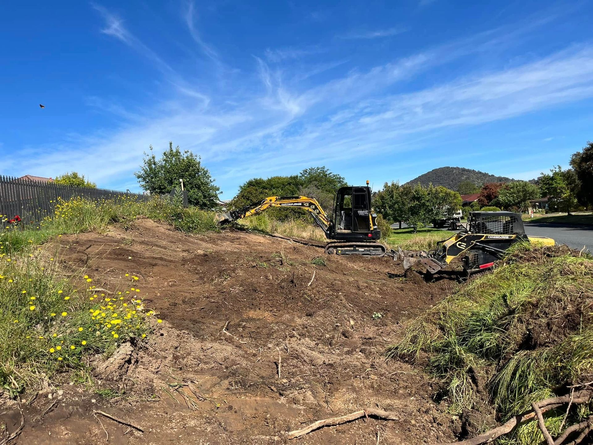 Bulldozers clearing dirt on a sunny hillside with trees and a blue sky
