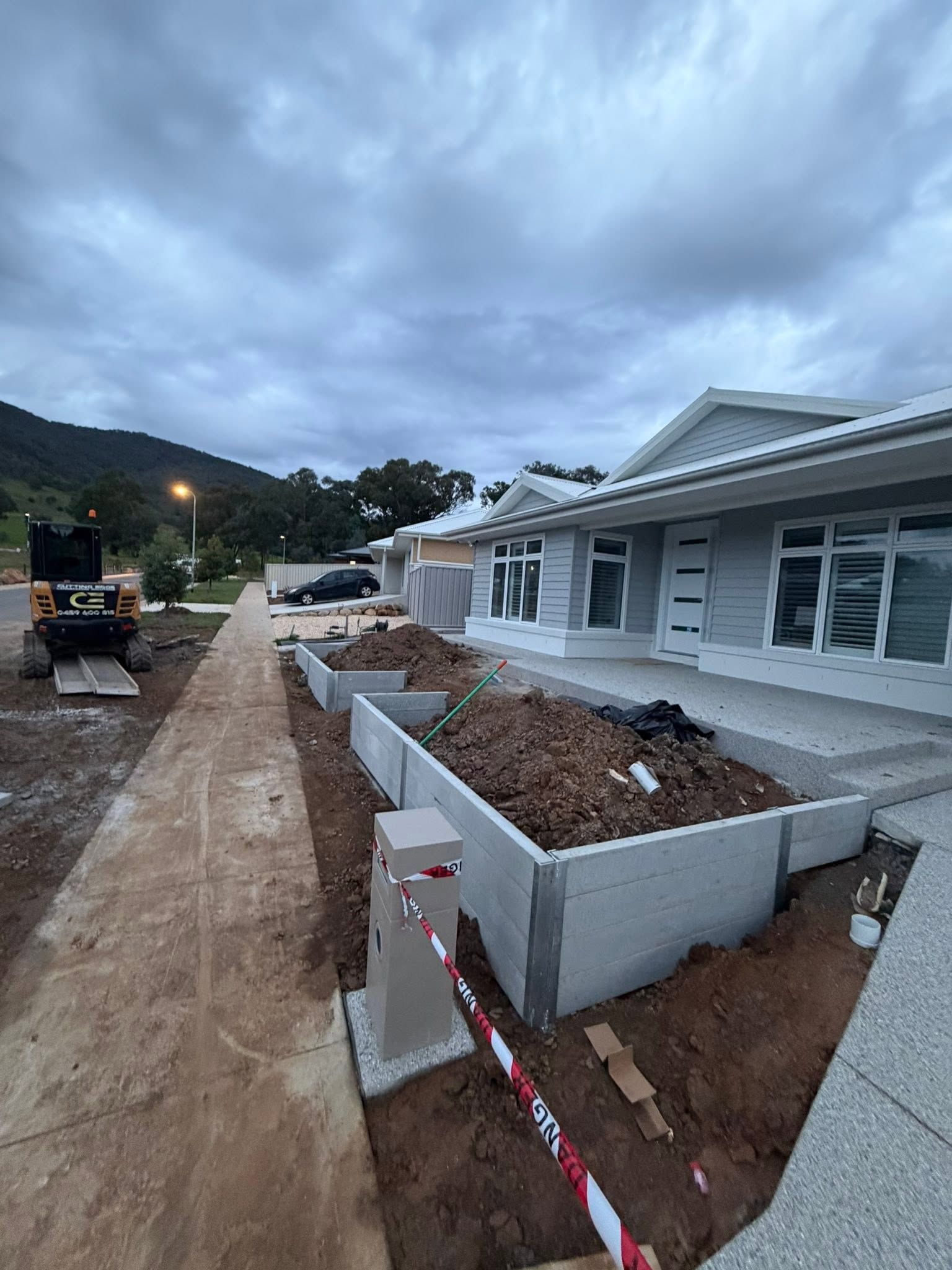 Construction site with raised concrete planters beside a house under a cloudy sky.