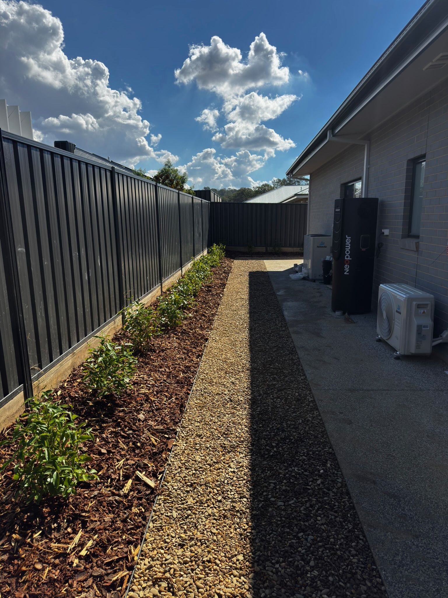 Narrow side yard with gravel path, mulch bed, black fence, and gray house wall under a blue sky