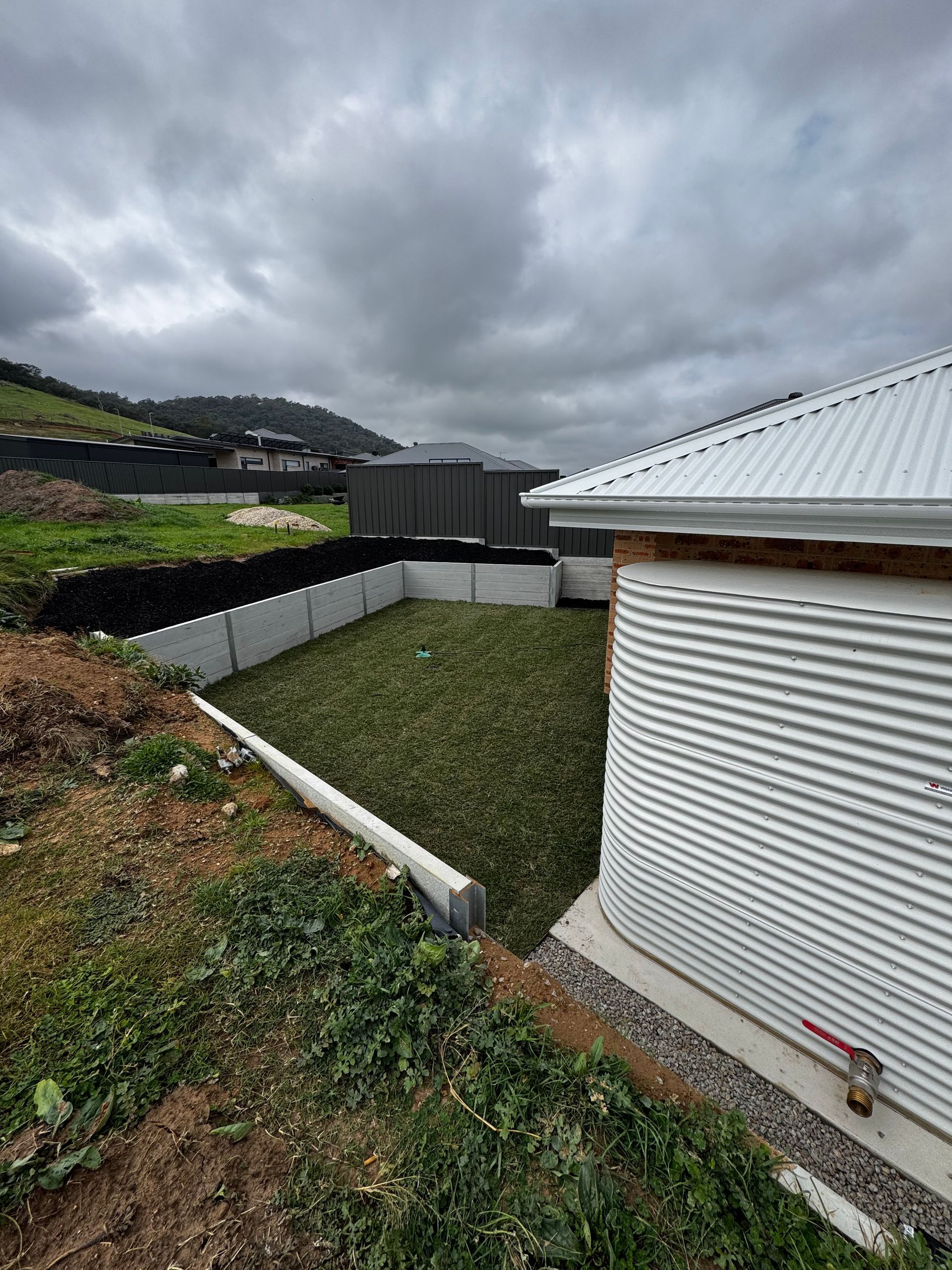 Overcast rural yard with a white corrugated building, grassy slope, and hillside homes in the distance