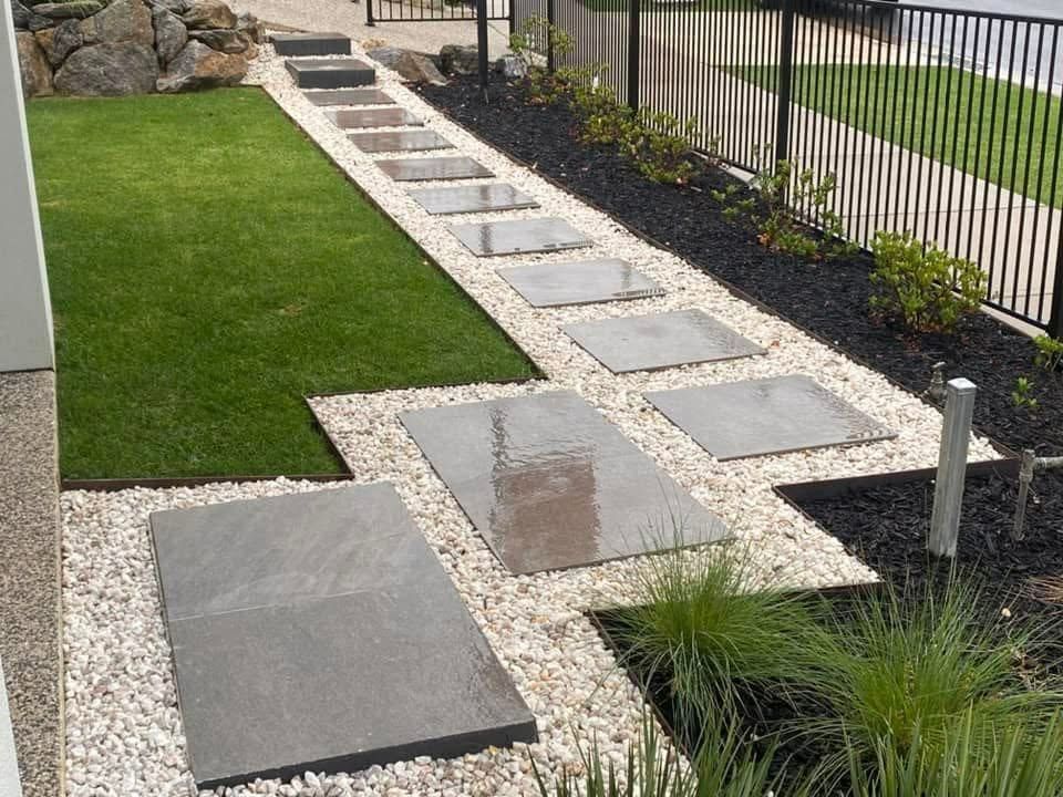 Modern stone walkway with square pavers beside a green lawn and black gravel border by a fence