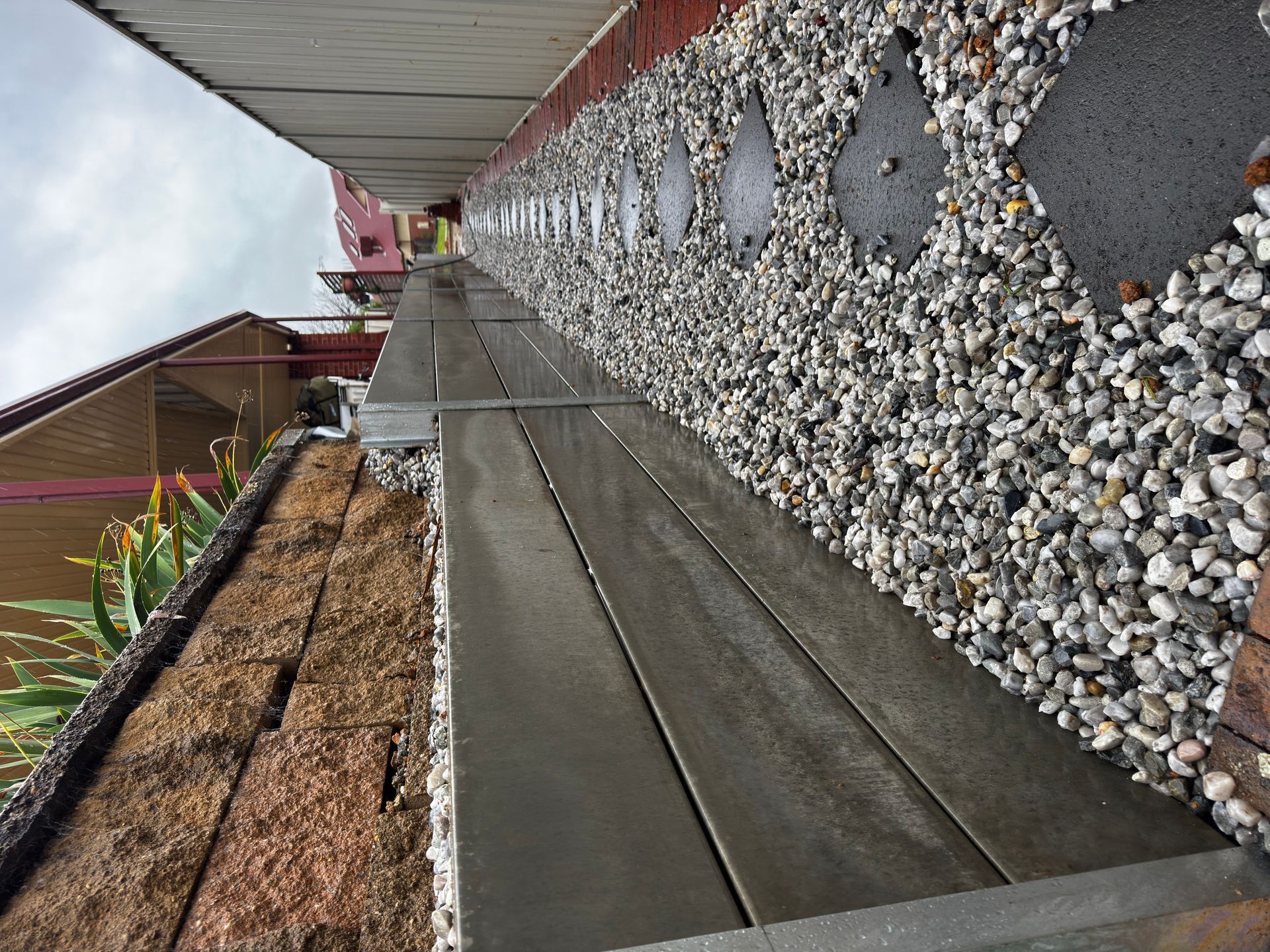 Long wooden walkway beside a stone wall with diamond accents and a flower bed on the left