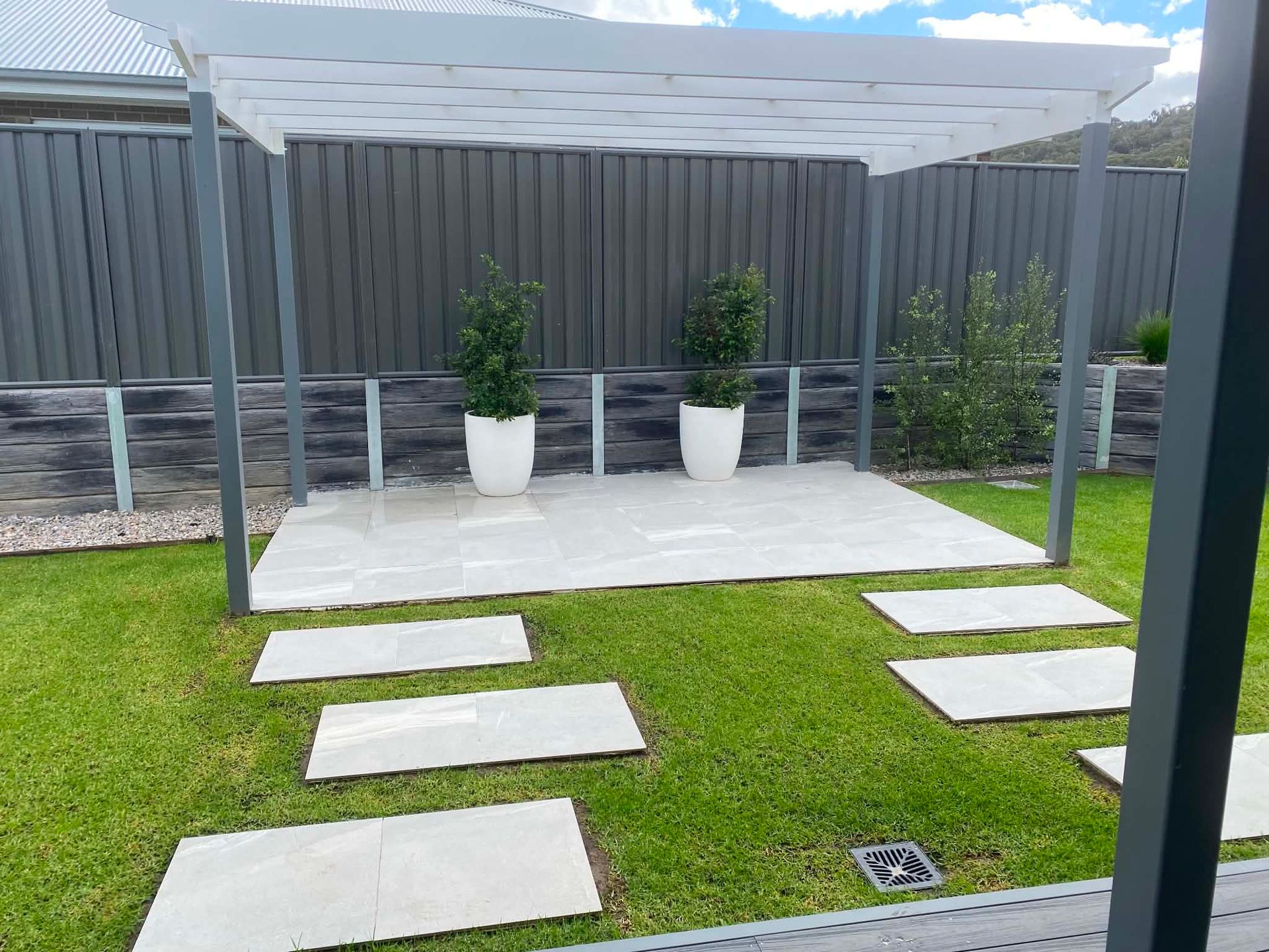 Backyard patio with white stepping stones, grass, potted plants, and a pergola beside a gray fence