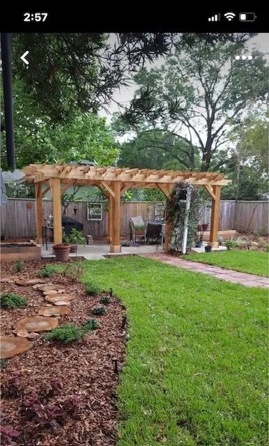 Wooden pergola in a backyard, with chairs and a walkway. Green grass and trees are in the background.